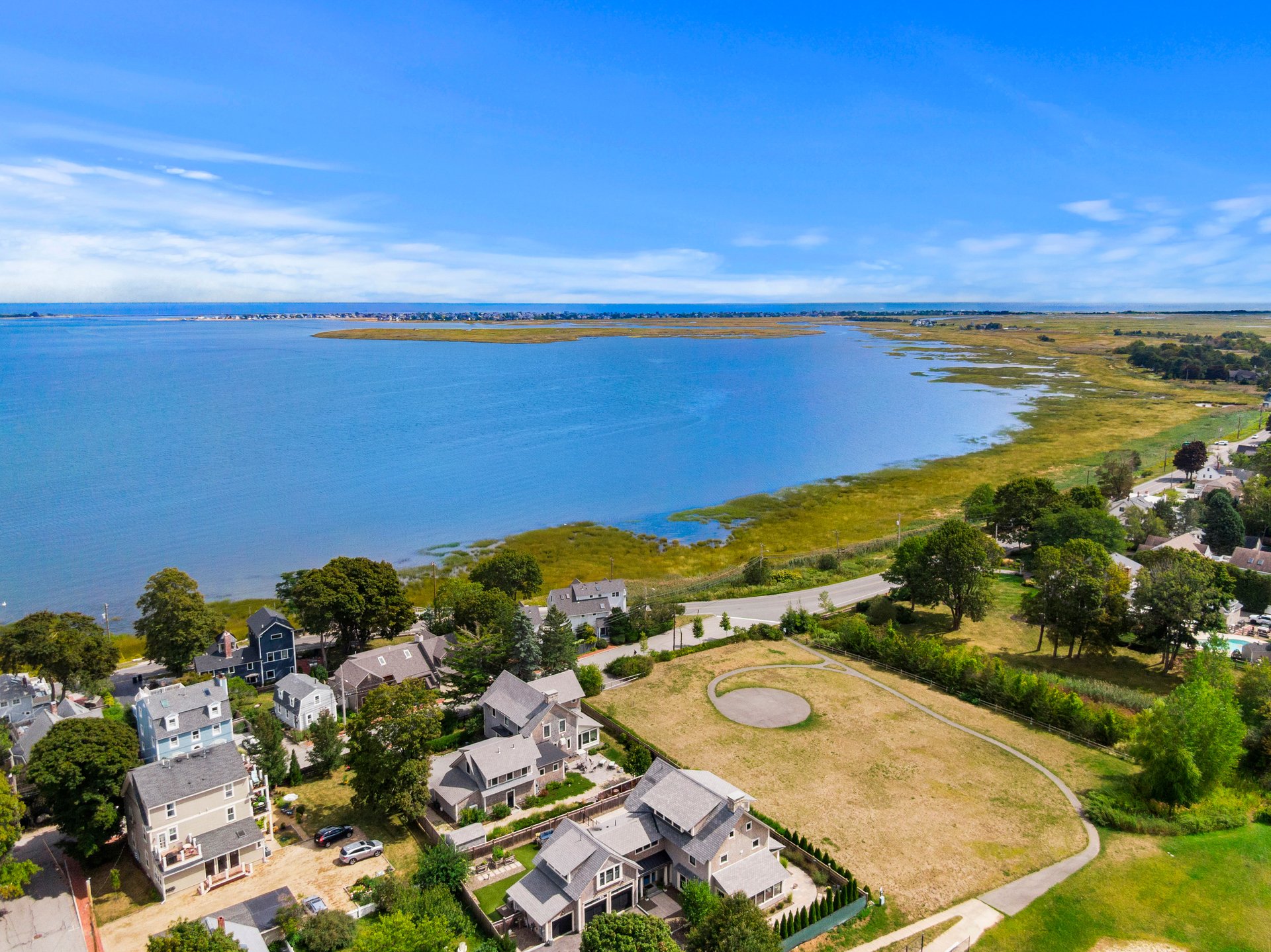 Aerial view of a coastal neighborhood, with houses beside a bay, marshland, and an open grassy lot.
