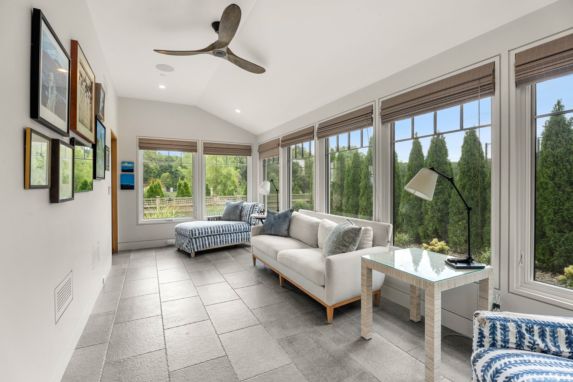 Furnished sunroom with broad garden-facing windows, a white sofa, and blue-and-white chaise lounges.