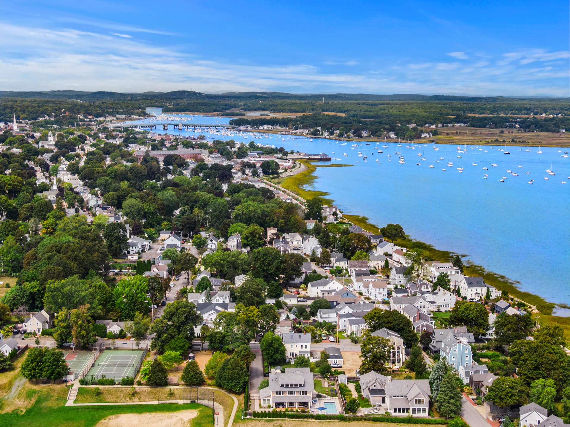 Aerial view of a coastal town and harbor, with sailboats, waterfront homes, and wooded hills.