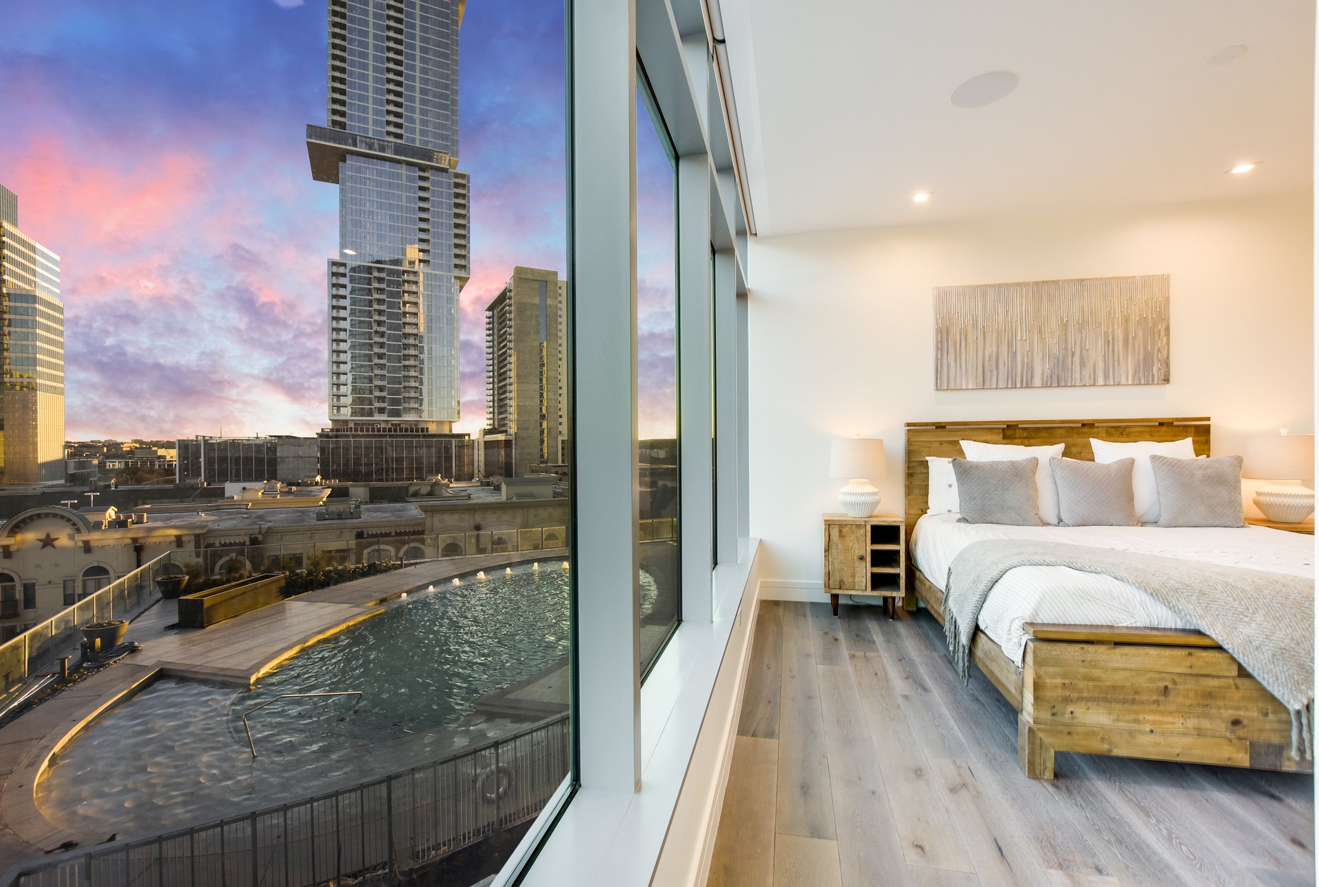 Modern condo bedroom overlooking a rooftop pool and downtown high-rises at sunset.