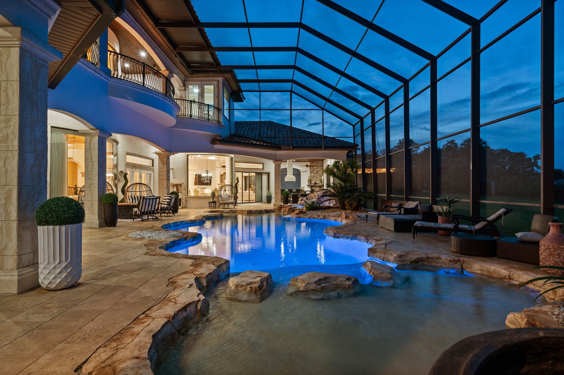 Screened pool and rock-edged spa beside a warmly lit two-story home at dusk