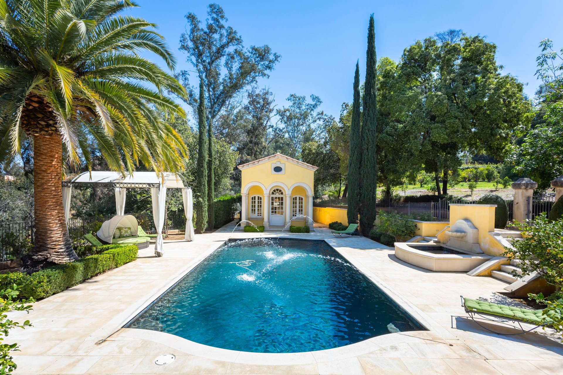 Estate swimming pool with a yellow arched pool house, stone fountain, and palm-shaded cabana.