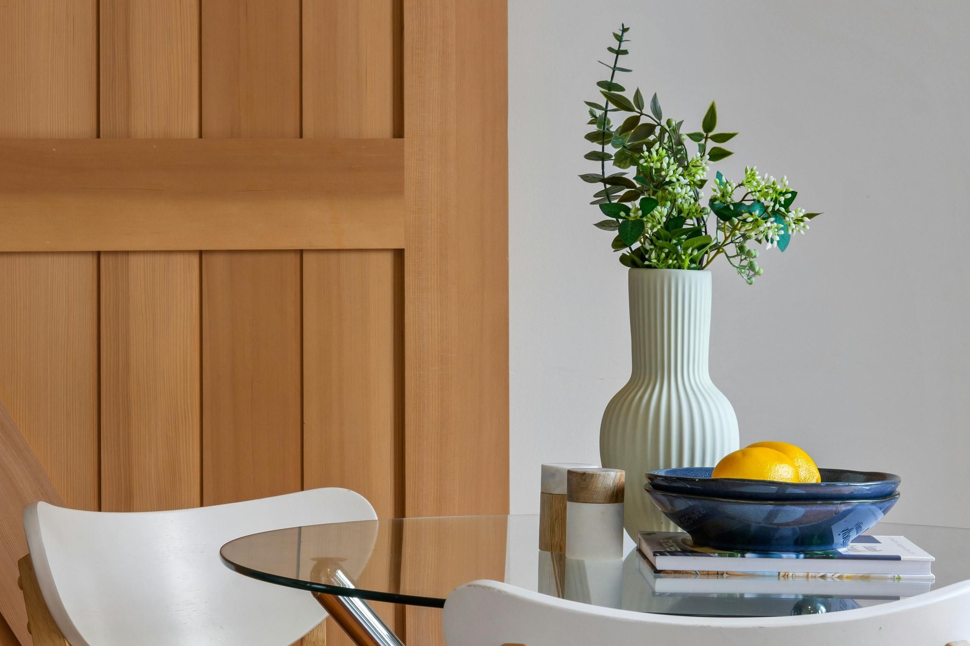 Dining area with a glass table, white chairs, warm wood paneling, and a vase of flowers.