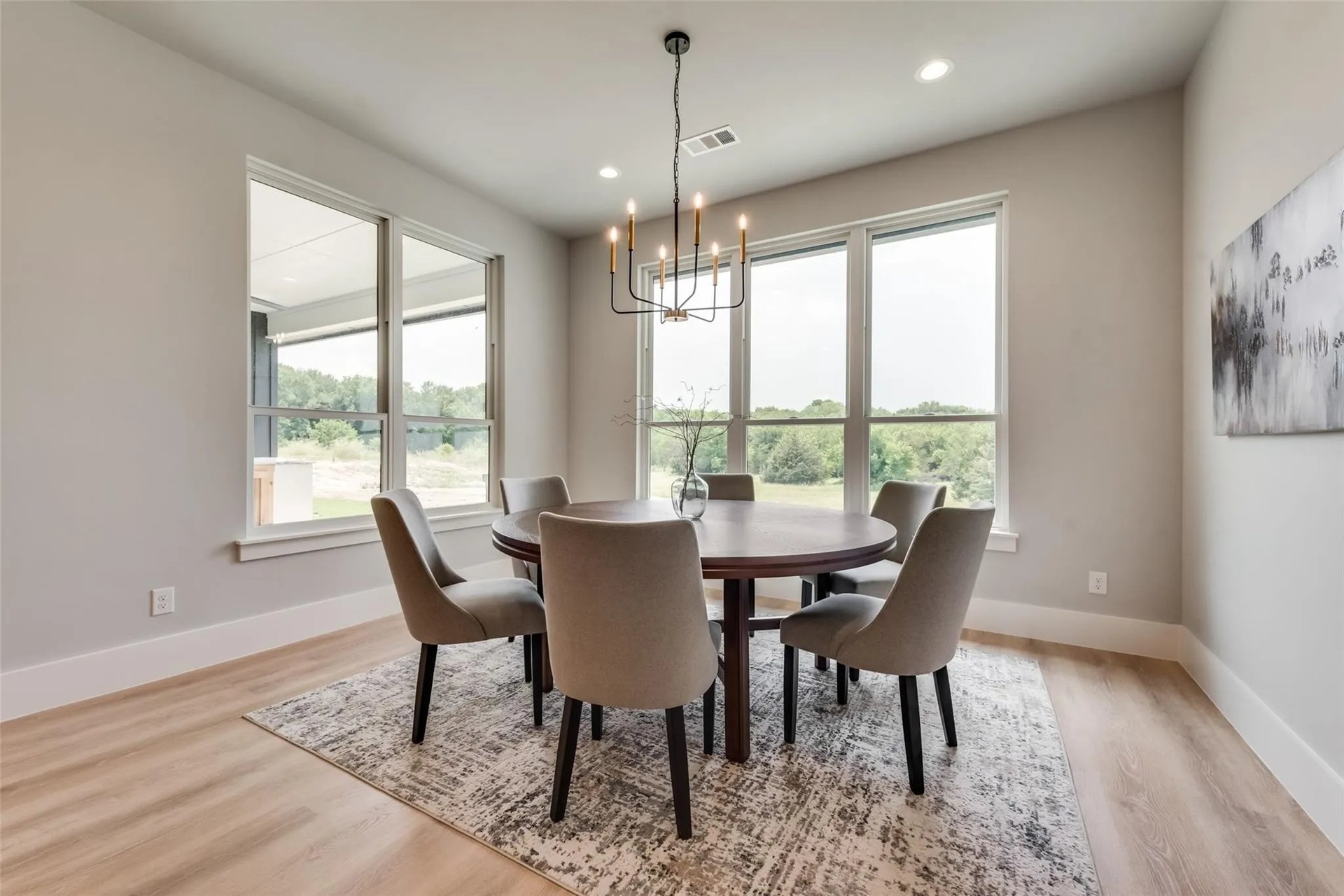 Dining room with a round table, upholstered chairs, modern chandelier, and large windows overlooking greenery.