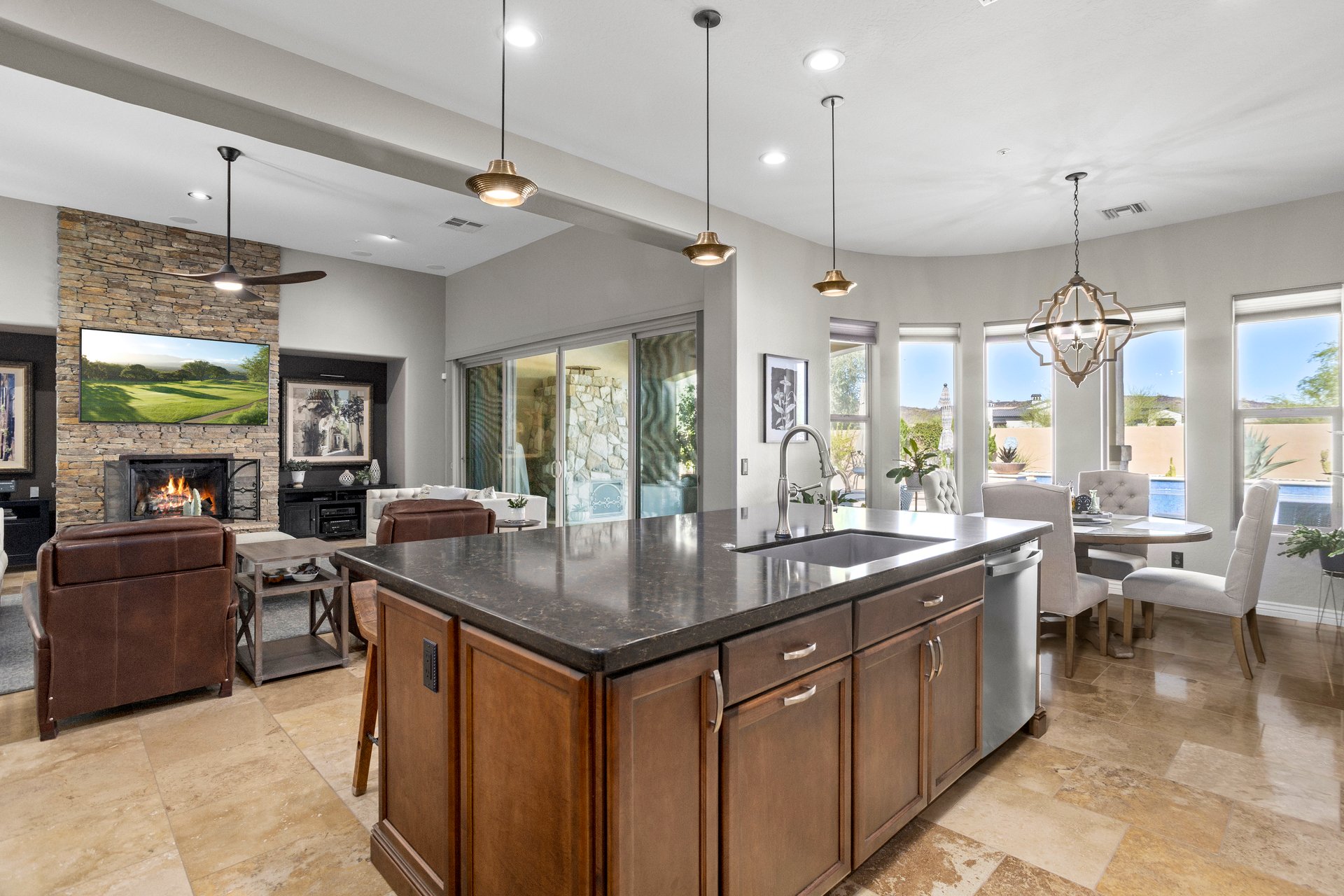 Open-concept kitchen with large island and sink, pendant lights, dining nook by curved windows, and a living area with stone fireplace in the background.