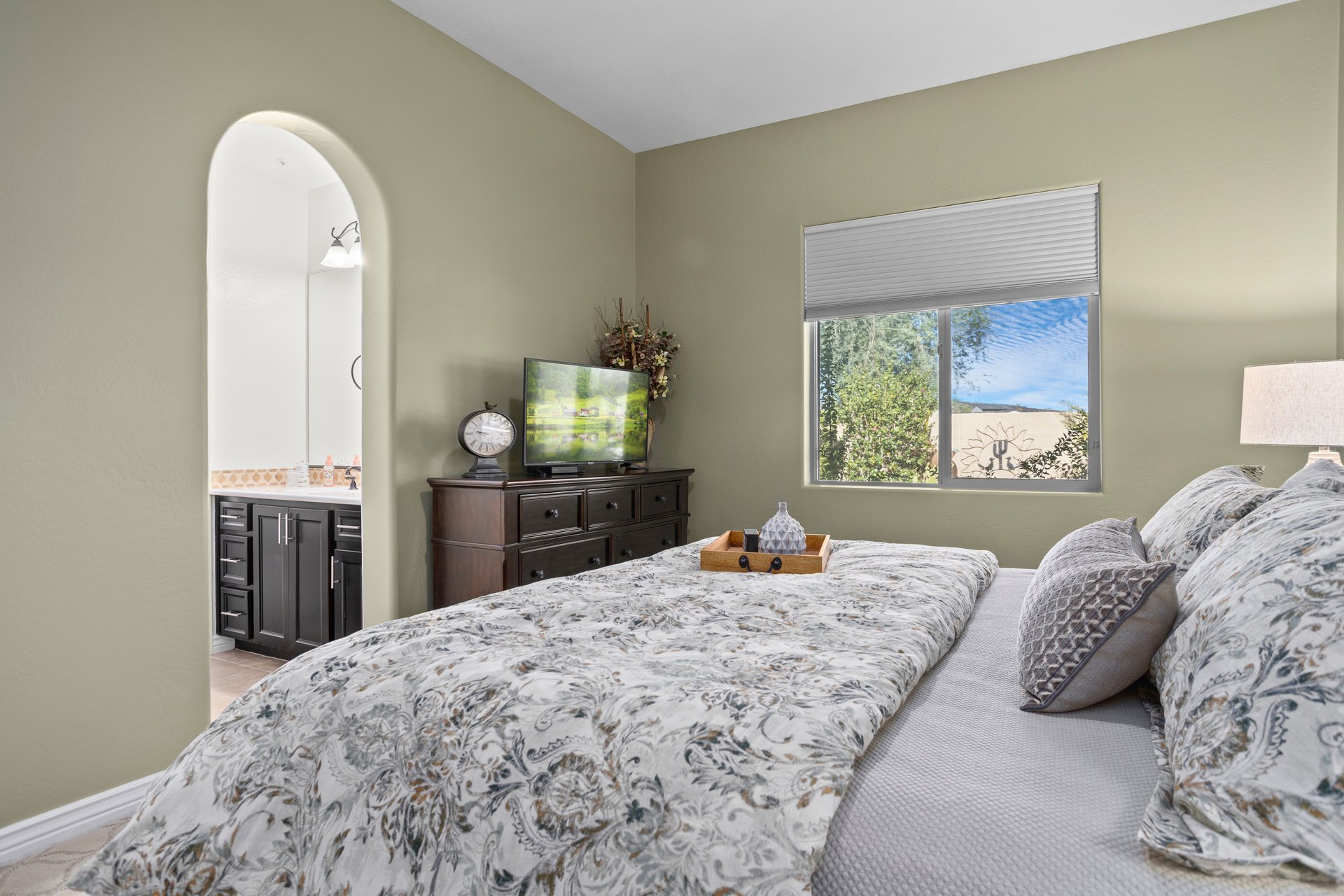 A furnished bedroom with a bed, dresser, window, and an arched opening leading to an ensuite bathroom vanity.