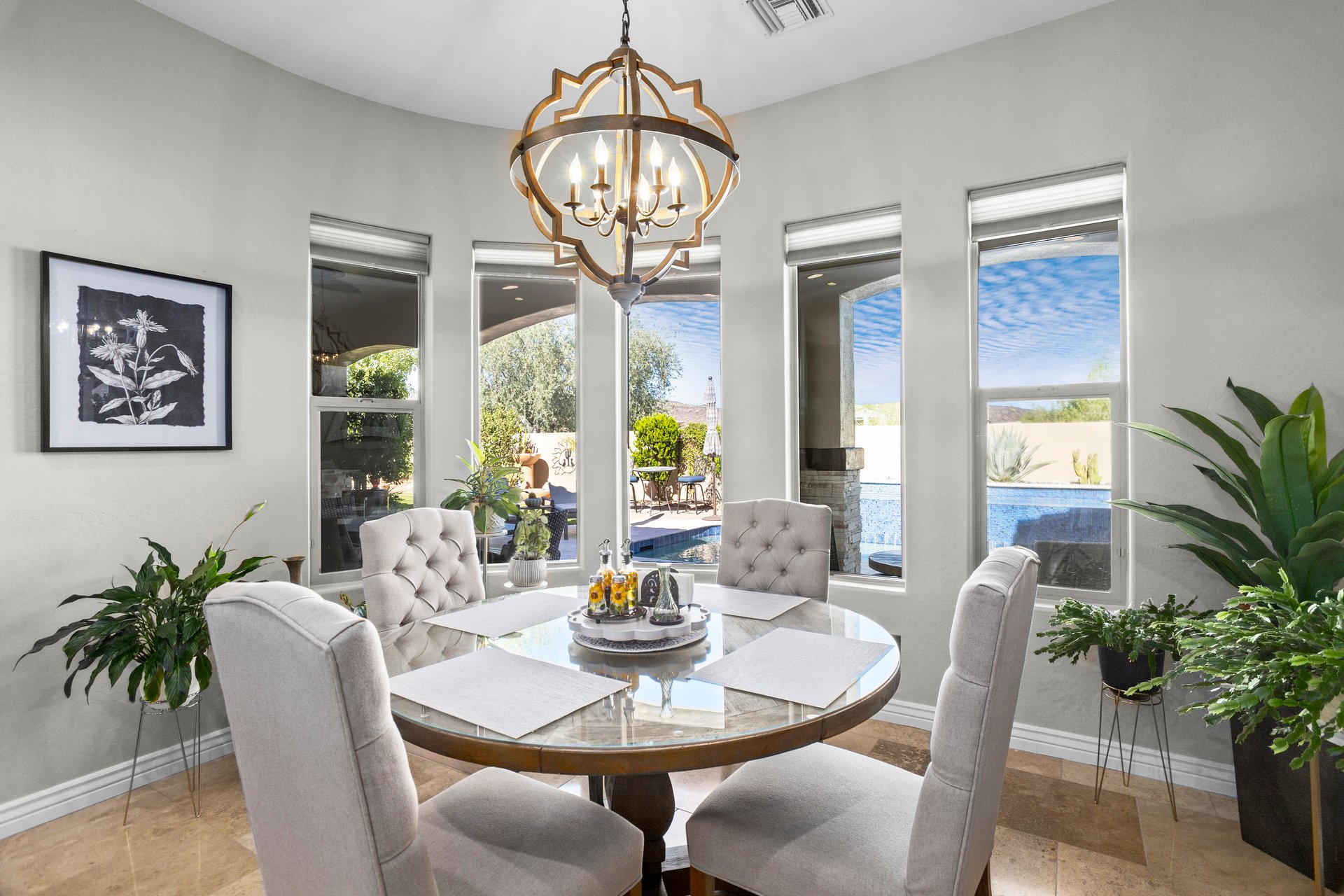 Staged dining room with a round glass table, upholstered chairs, chandelier, and tall windows looking out to a backyard patio and pool.