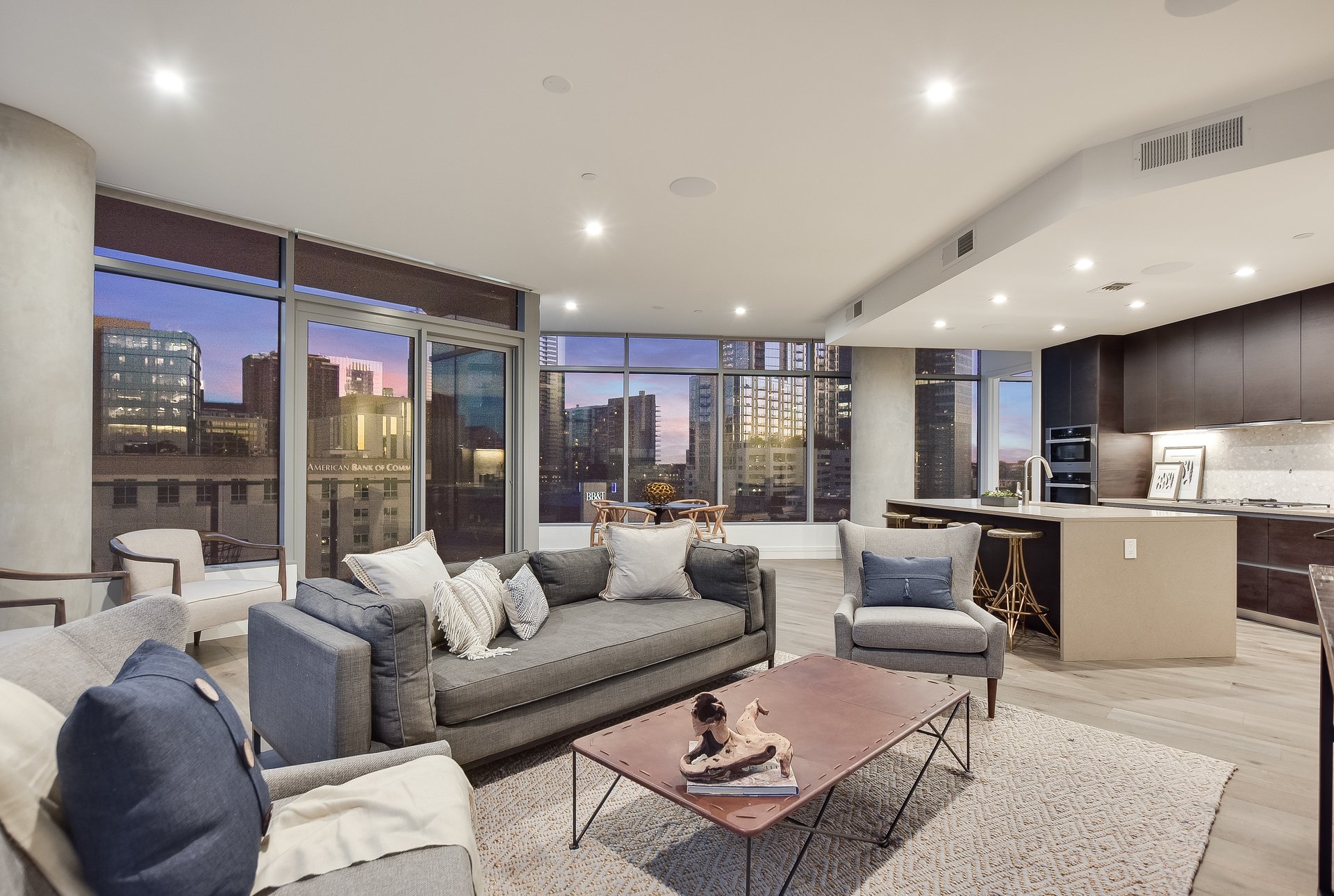 Furnished condo living room with floor-to-ceiling windows overlooking the downtown skyline at dusk.