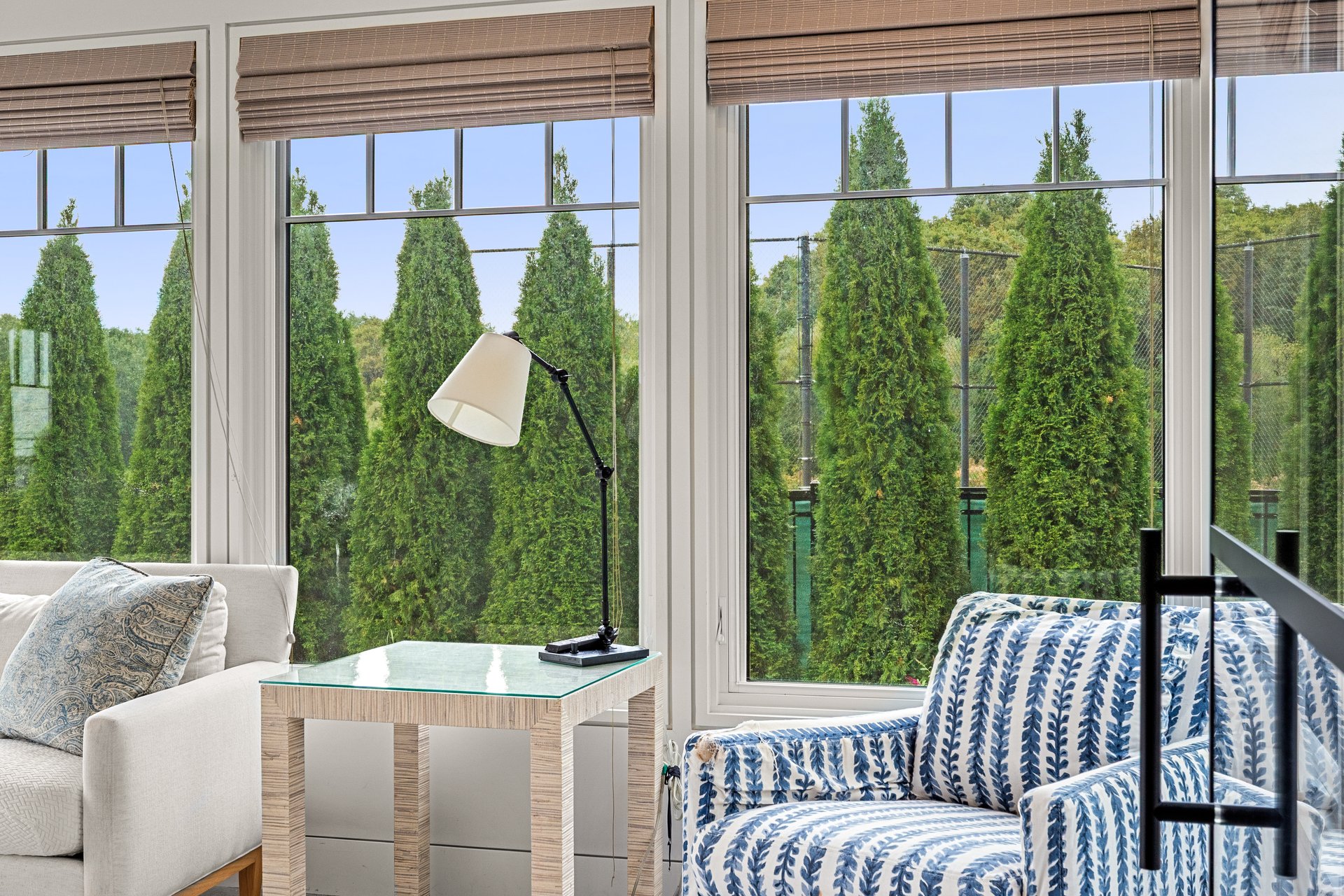 Furnished sitting area with patterned seating, a glass-topped table, and large windows overlooking evergreens.