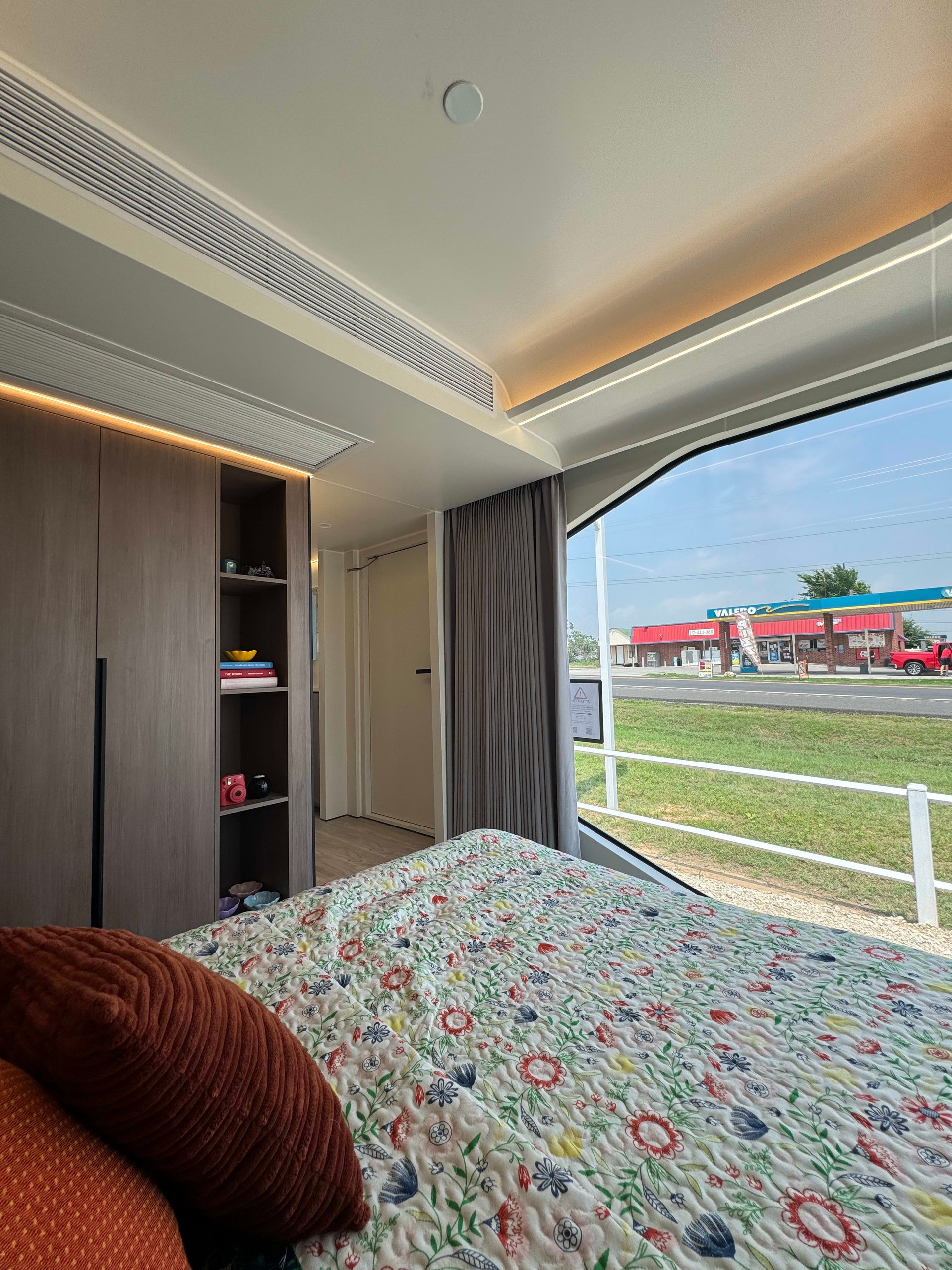 Tiny home bedroom with a floral quilted bed, built-in shelving, and a large curved window.