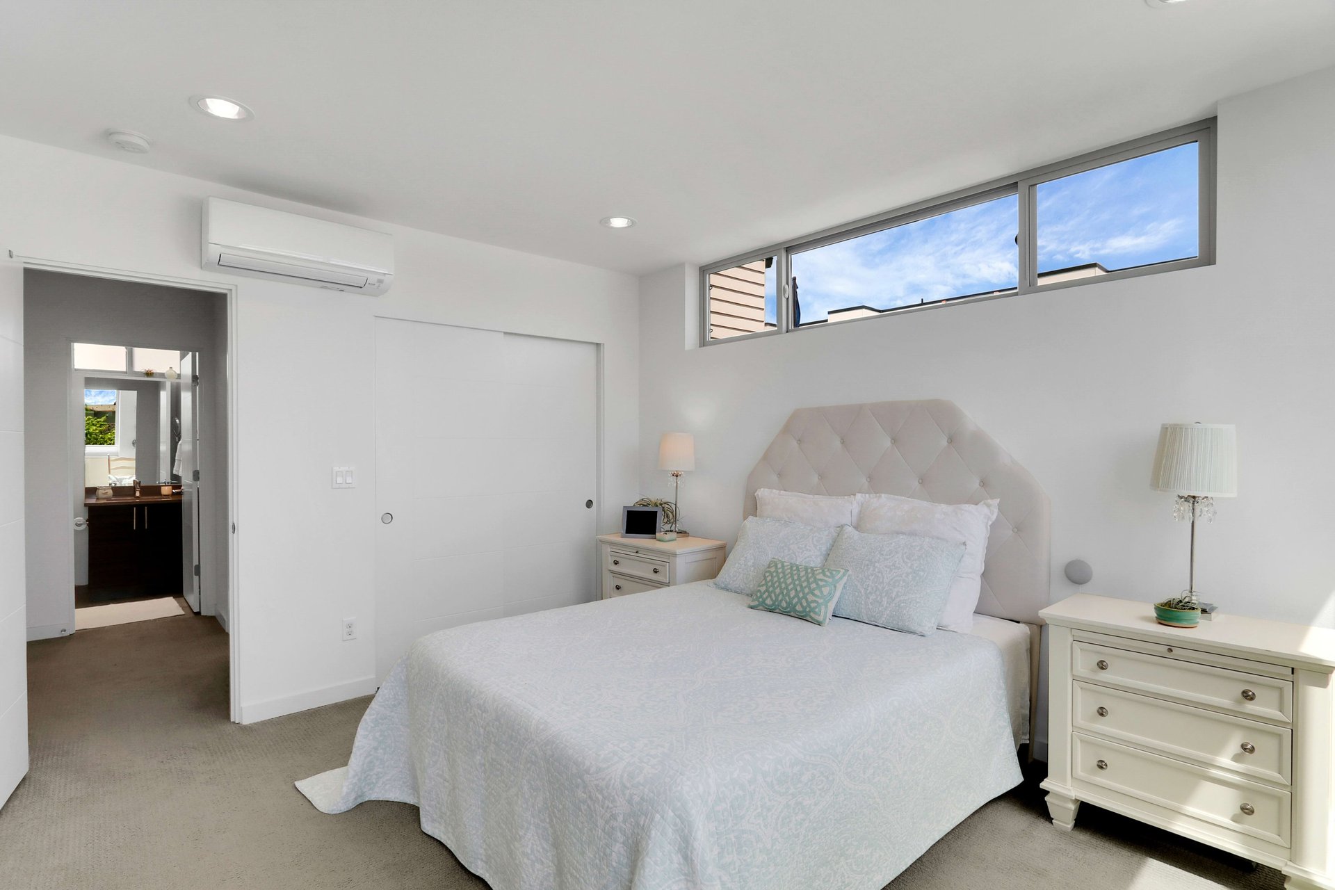 Bright staged bedroom with a tufted headboard, high clerestory windows, and an adjoining bathroom doorway.