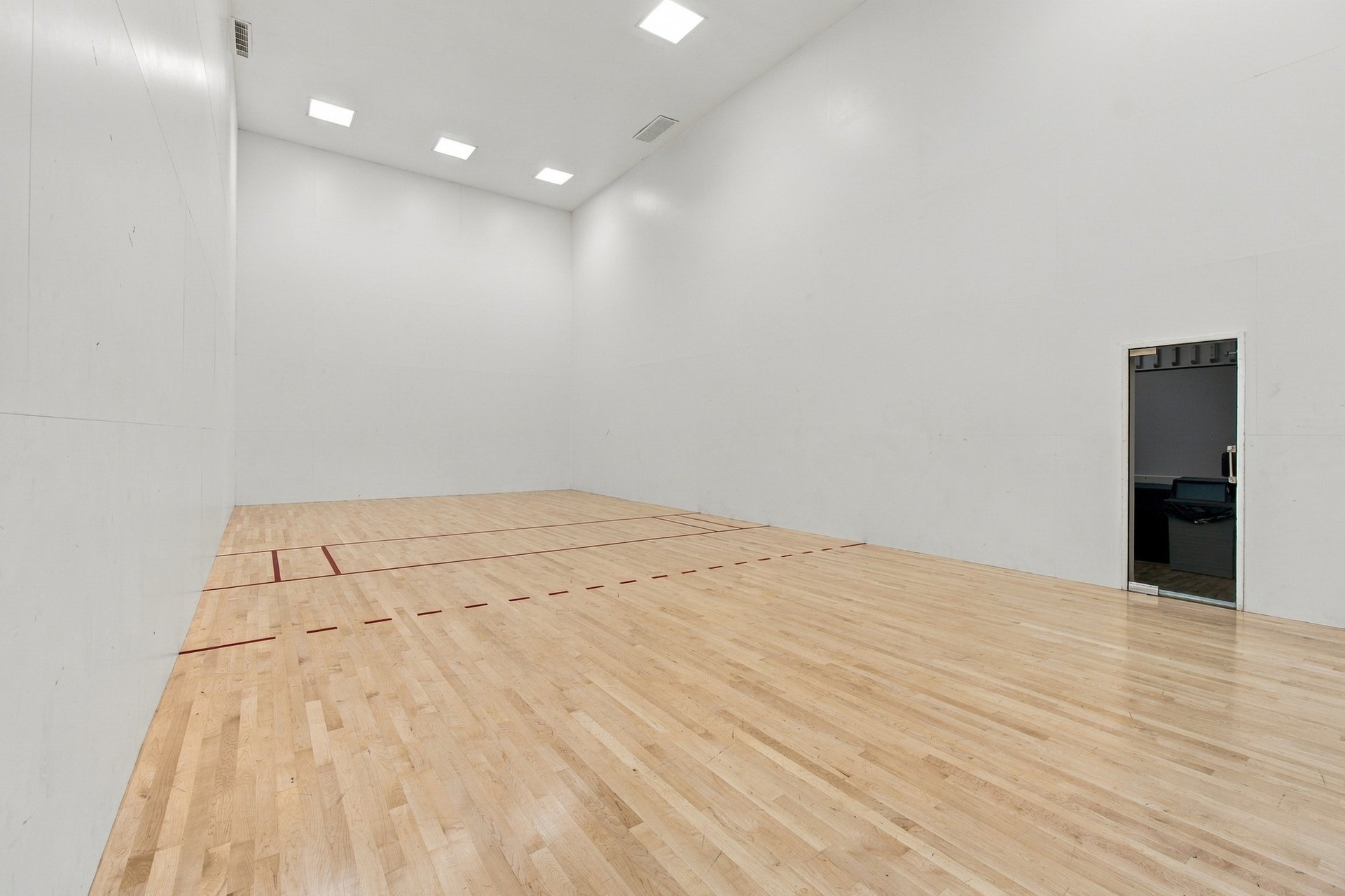 Empty squash court with red floor markings, pale wood flooring, and tall white walls.