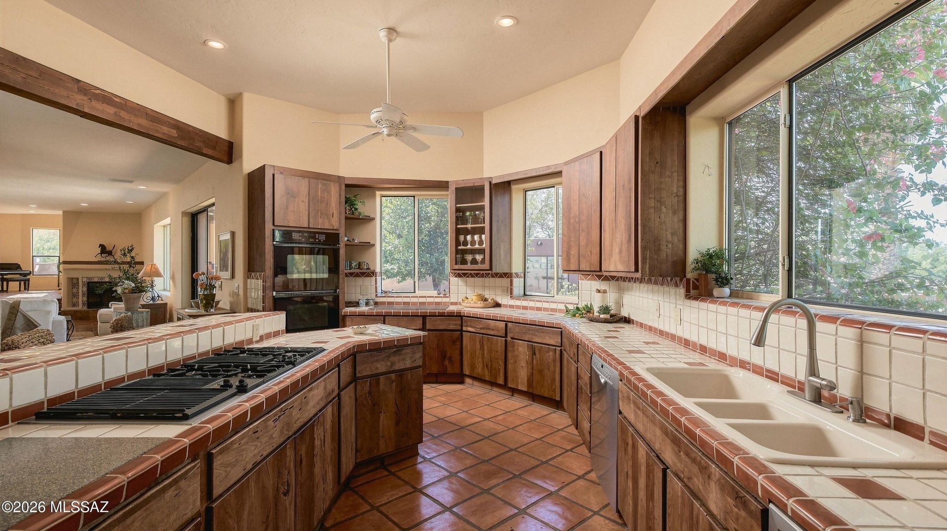Kitchen with dark wood cabinets, tiled counters, gas cooktop, double ovens, and large windows overlooking trees.