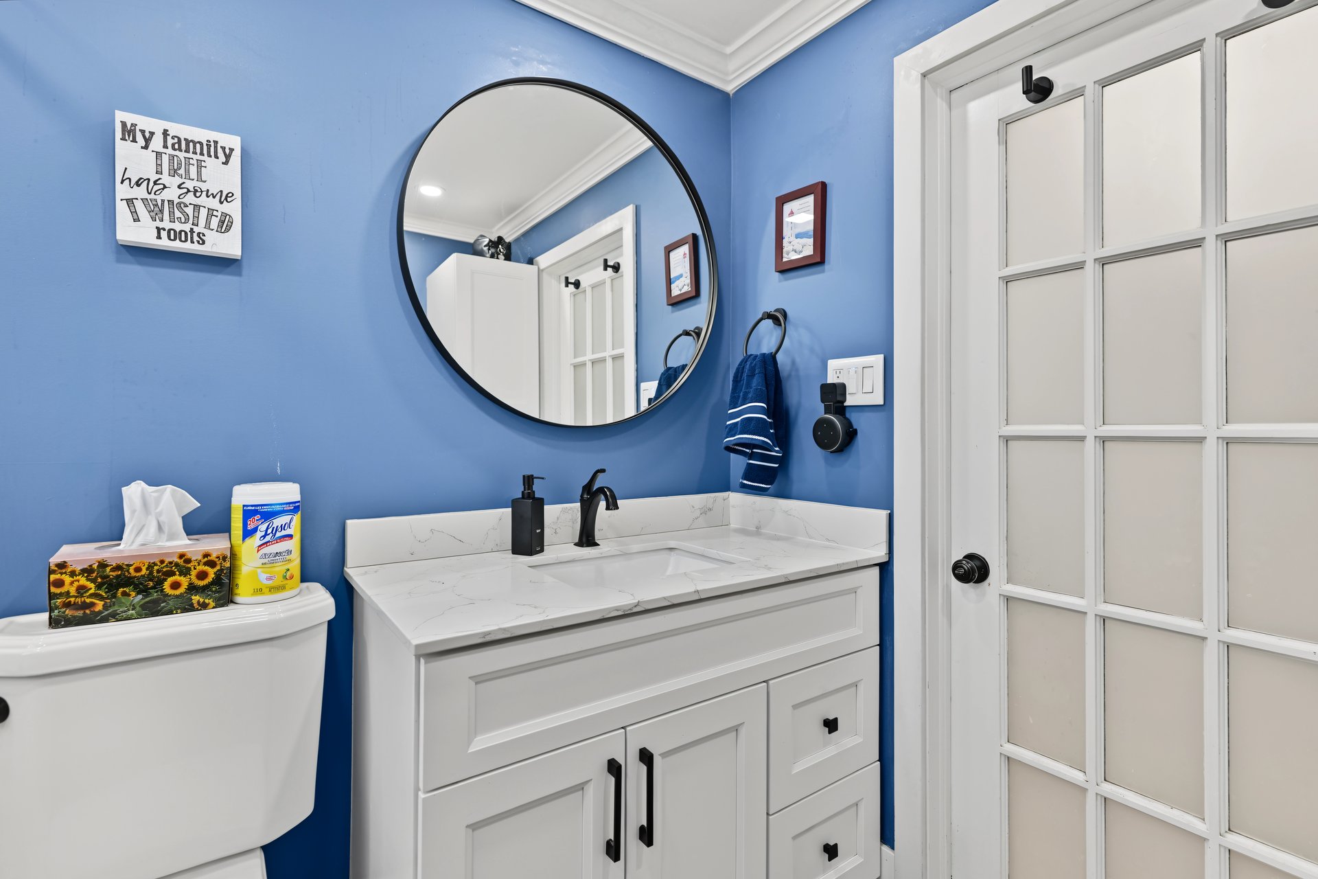 Bathroom with blue walls, white vanity, marble-look countertop, round mirror, and glass-paneled door.