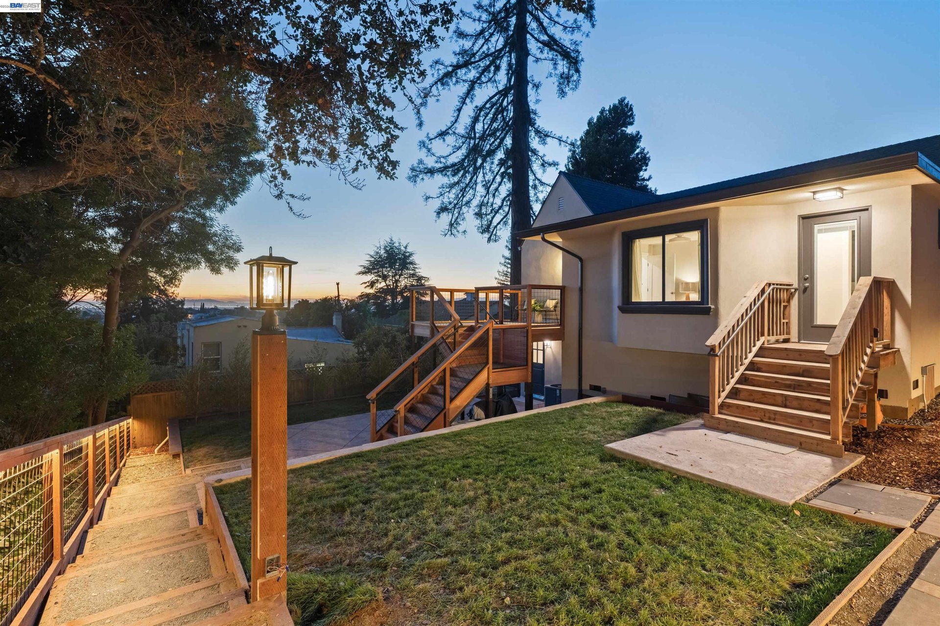 Oakland home at dusk with a raised wooden deck, fenced yard, and illuminated path lamp.