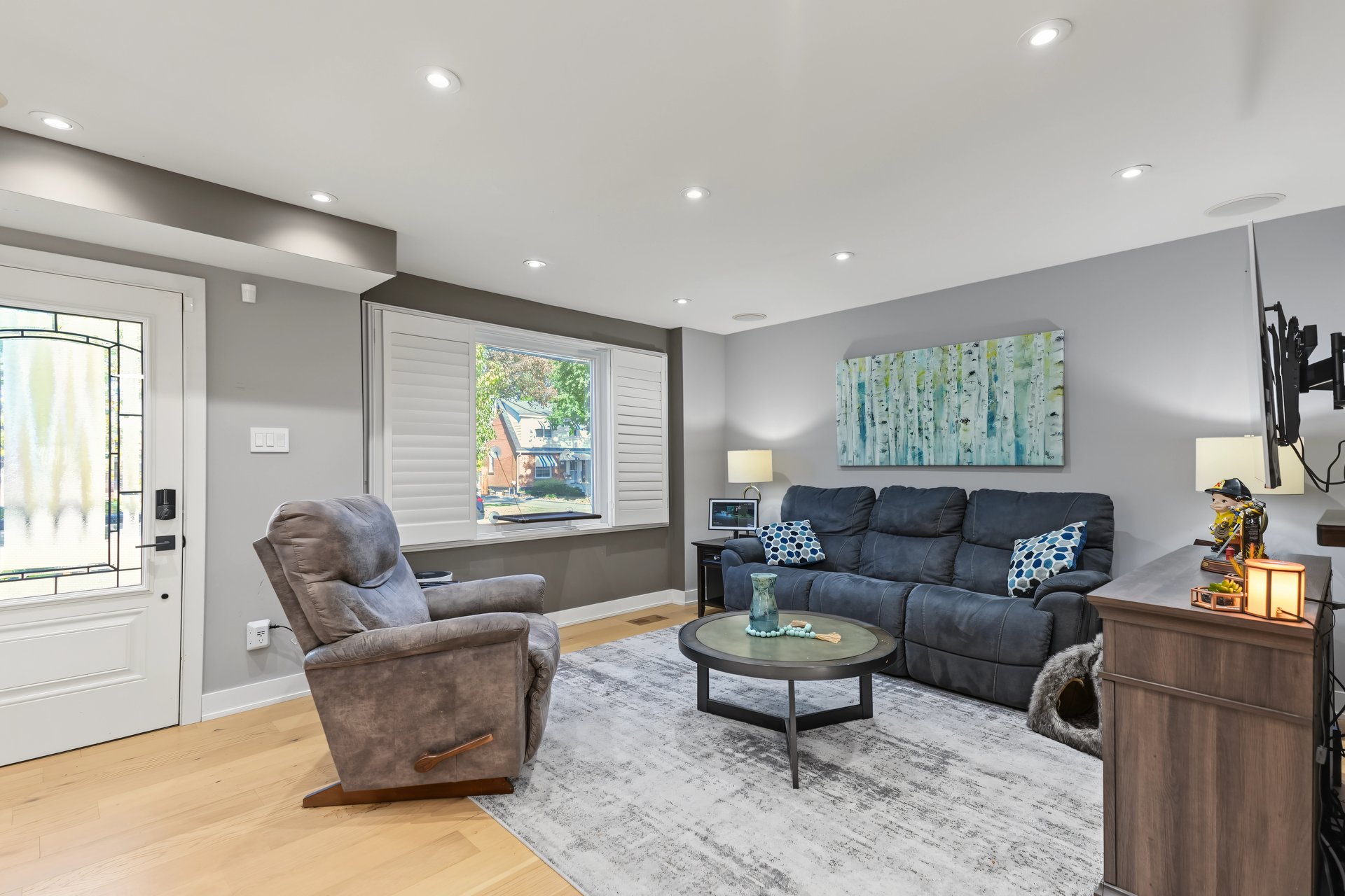 Furnished living room with a dark sectional, gray recliner, and white-shuttered window.