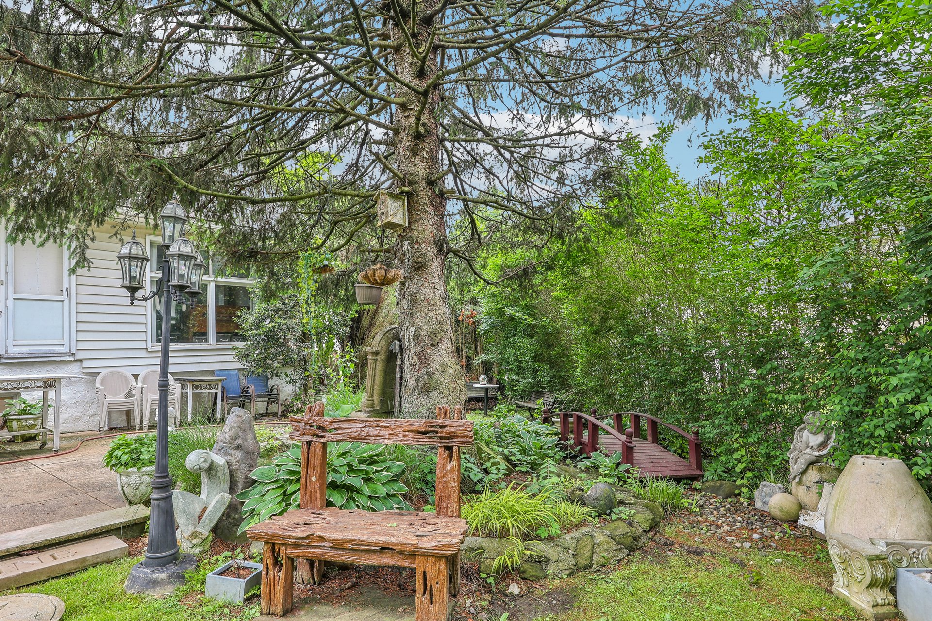 Landscaped backyard with a large evergreen, rustic bench, garden ornaments, and a small red footbridge.