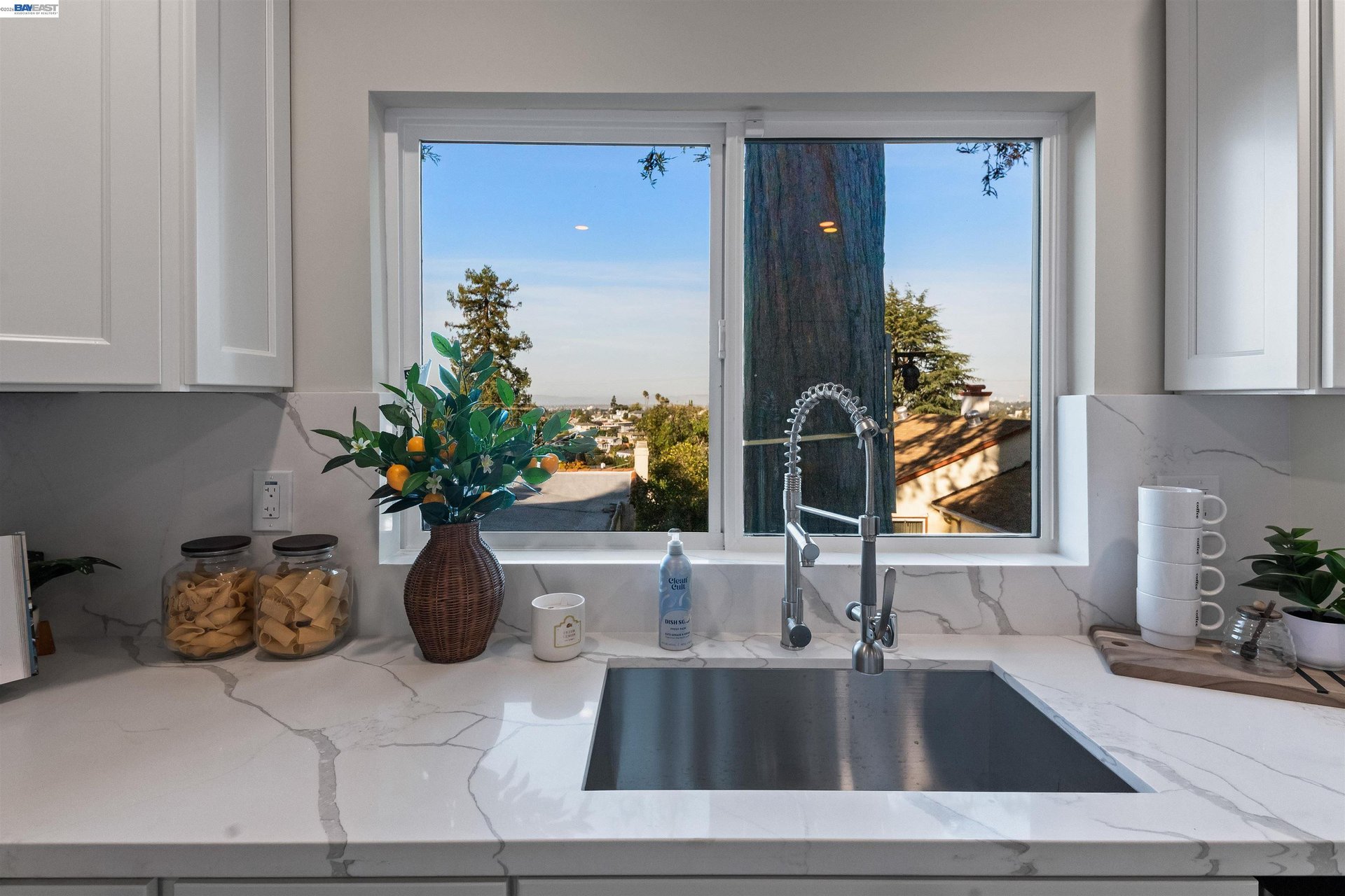 Kitchen sink beneath a wide window overlooking nearby homes, with white veined counters and a tall spring-coil faucet.