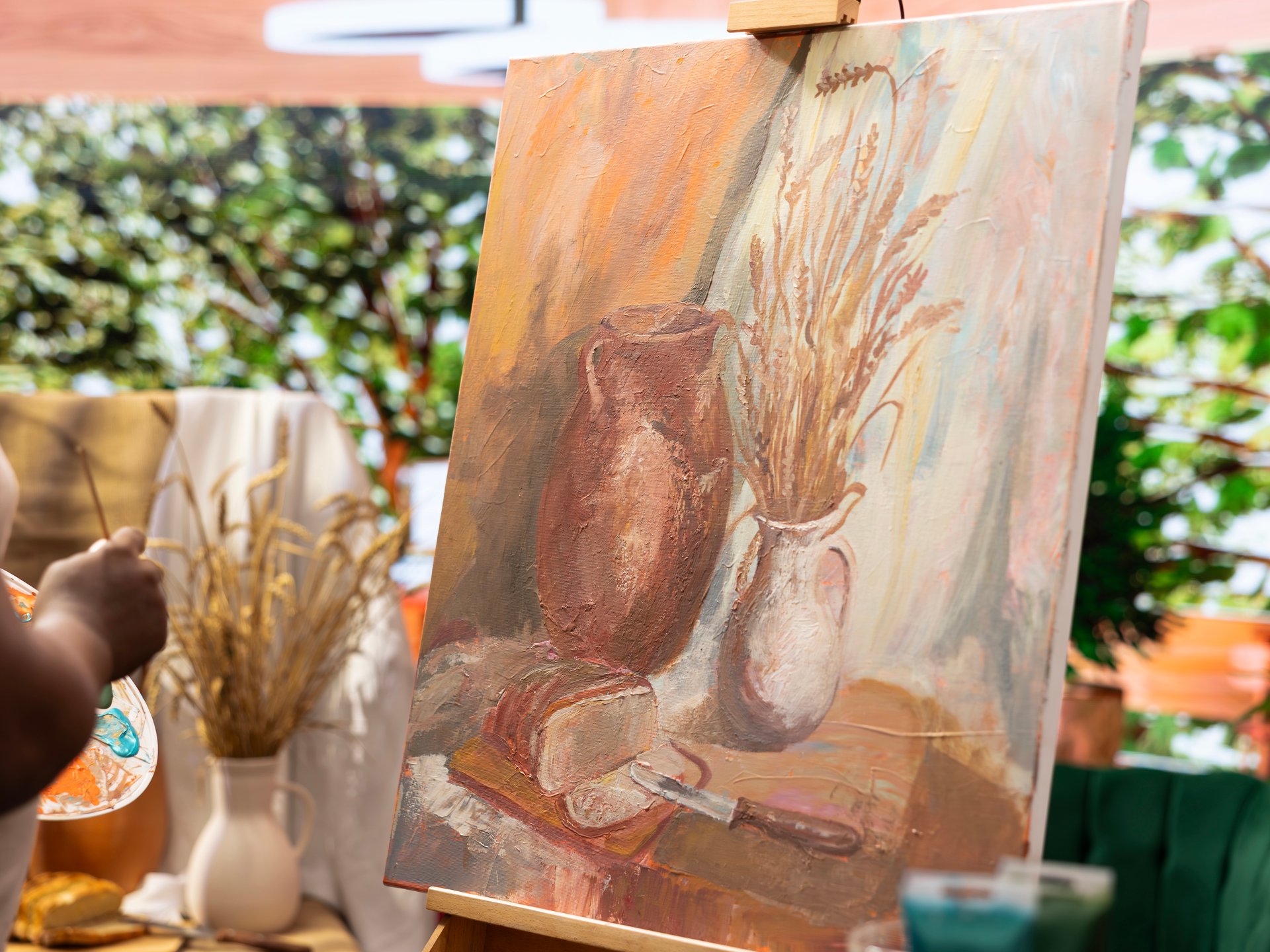 Still-life painting on an easel at an outdoor art market, with the artist's palette and hand at left.