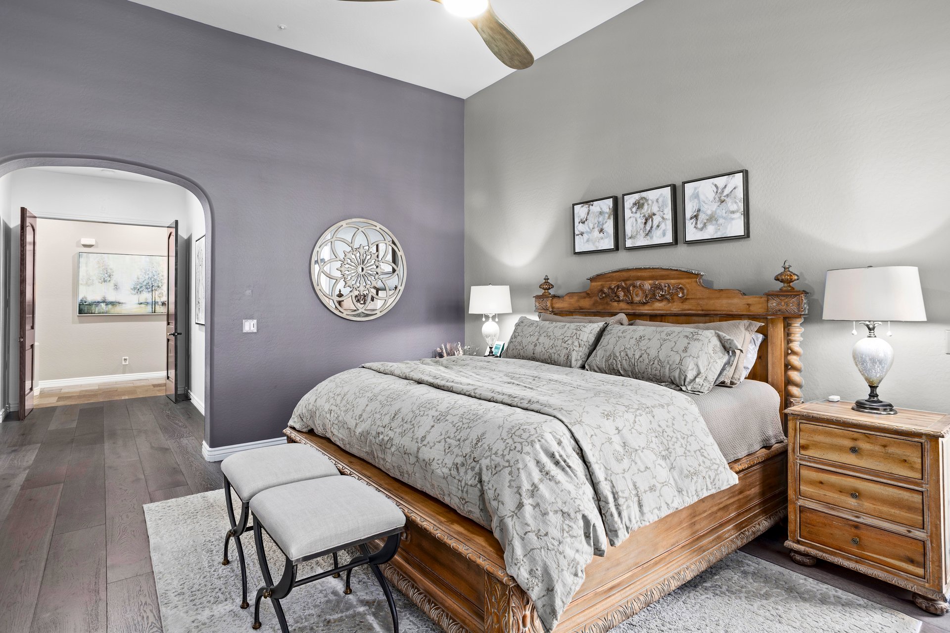 A furnished bedroom with a carved wooden bed, matching nightstands, lamps, artwork above the headboard, and an arched doorway leading to another room.