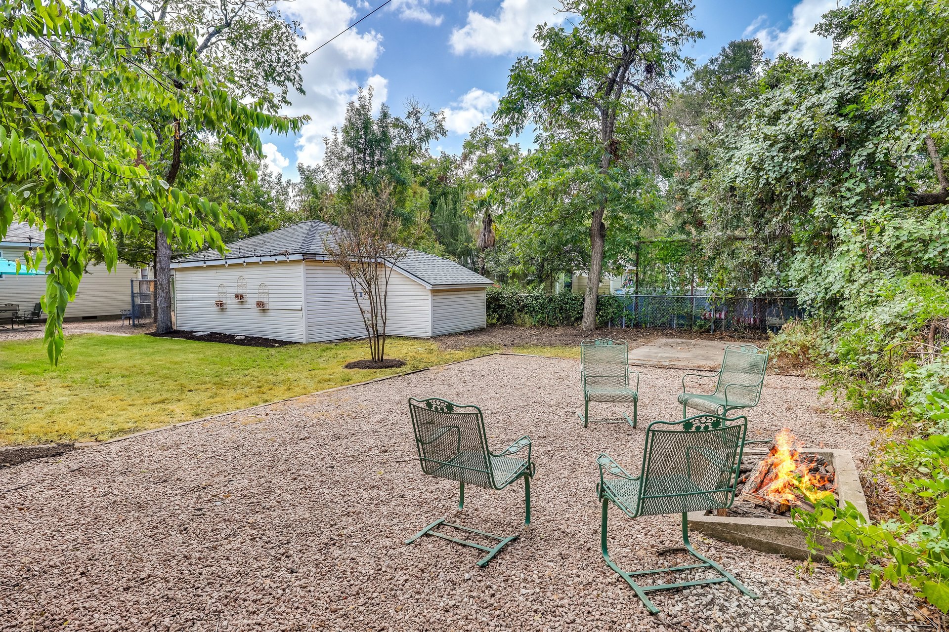 Backyard gravel patio with metal chairs around a fire pit beside a white outbuilding and mature trees.