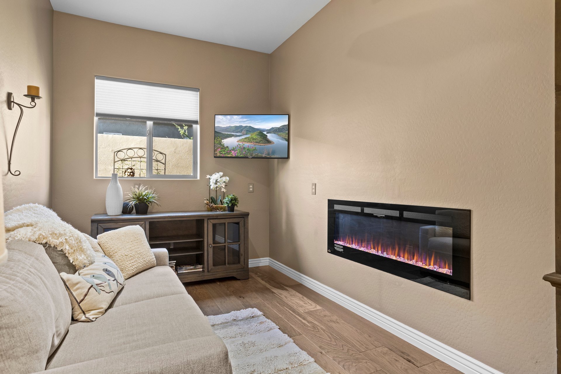 Staged living room with a sofa, wall-mounted electric fireplace, window, and console table with decor.