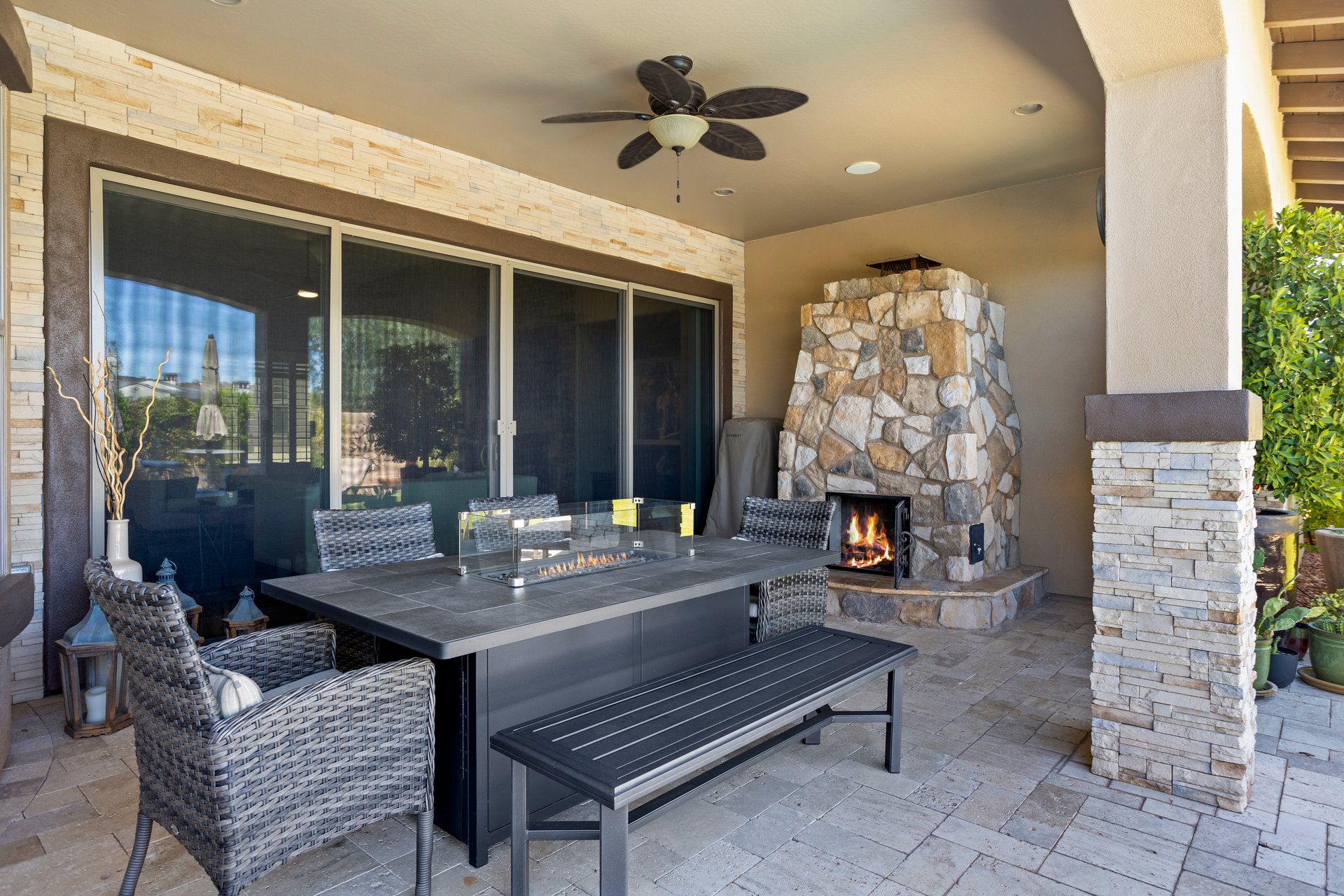 Covered outdoor patio with stone fireplace, dining table with built-in fire feature, and sliding glass doors to the house.
