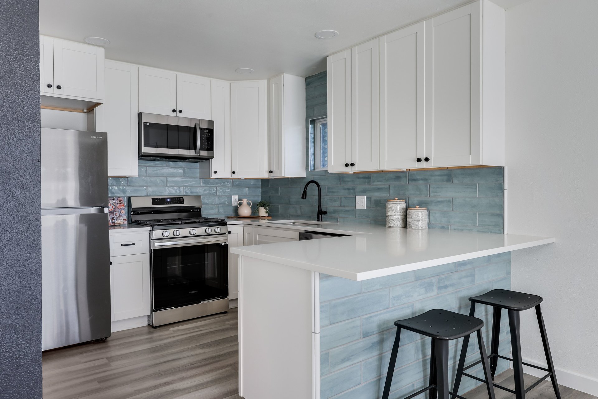 Kitchen with white cabinets, blue-gray tile backsplash, and a large peninsula with two black stools.