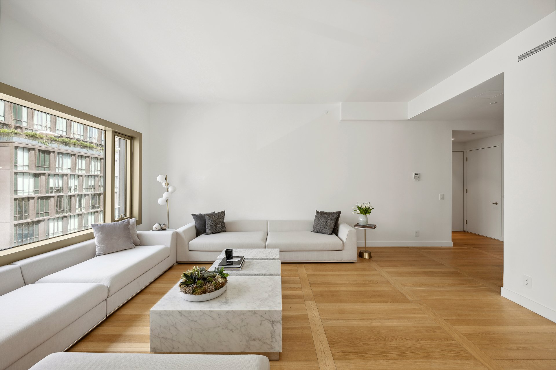 Living room with white sectional sofas, a marble coffee table, and large windows overlooking a neighboring building.