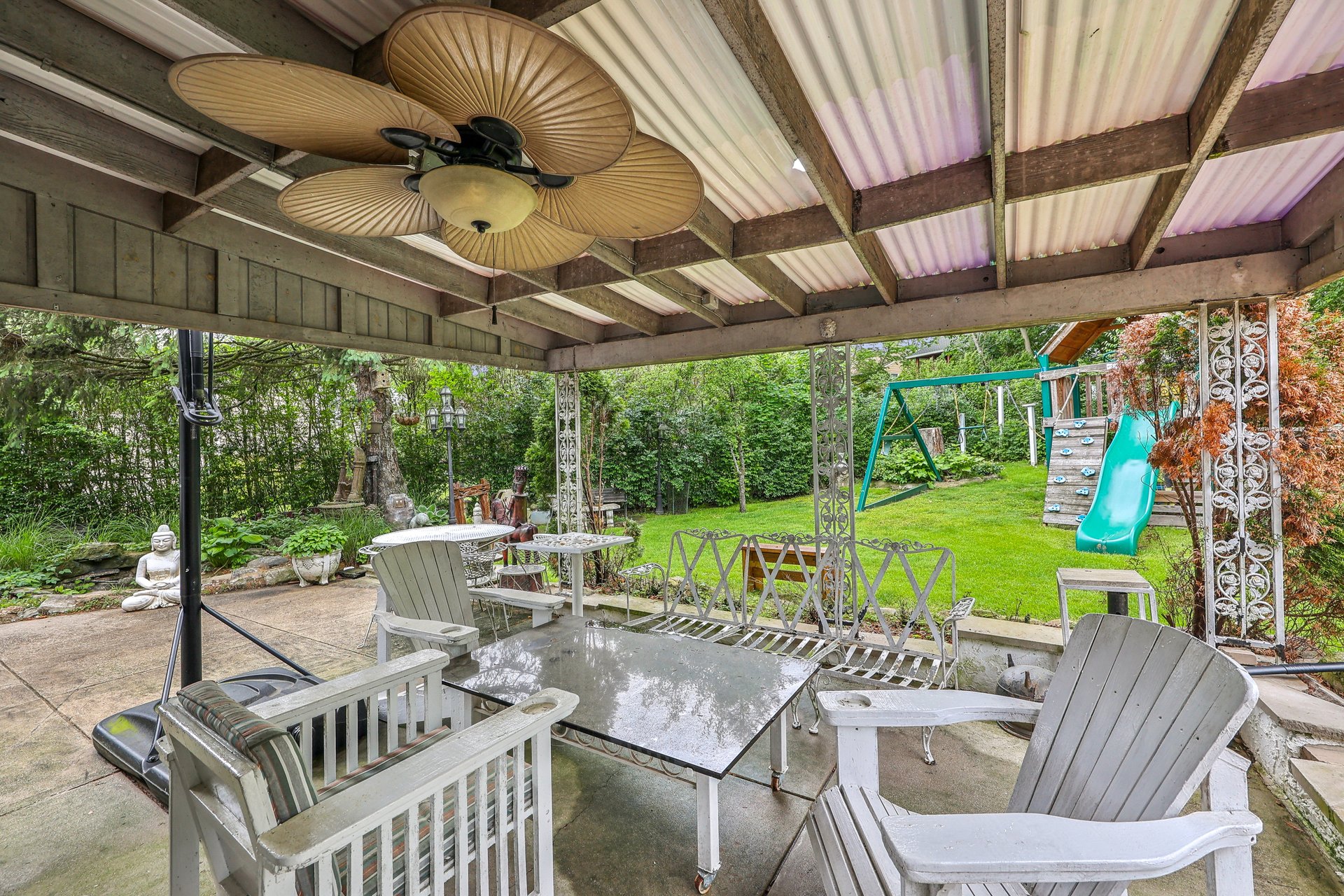 Covered patio with white outdoor seating and a ceiling fan, overlooking a lawn and children's playset.