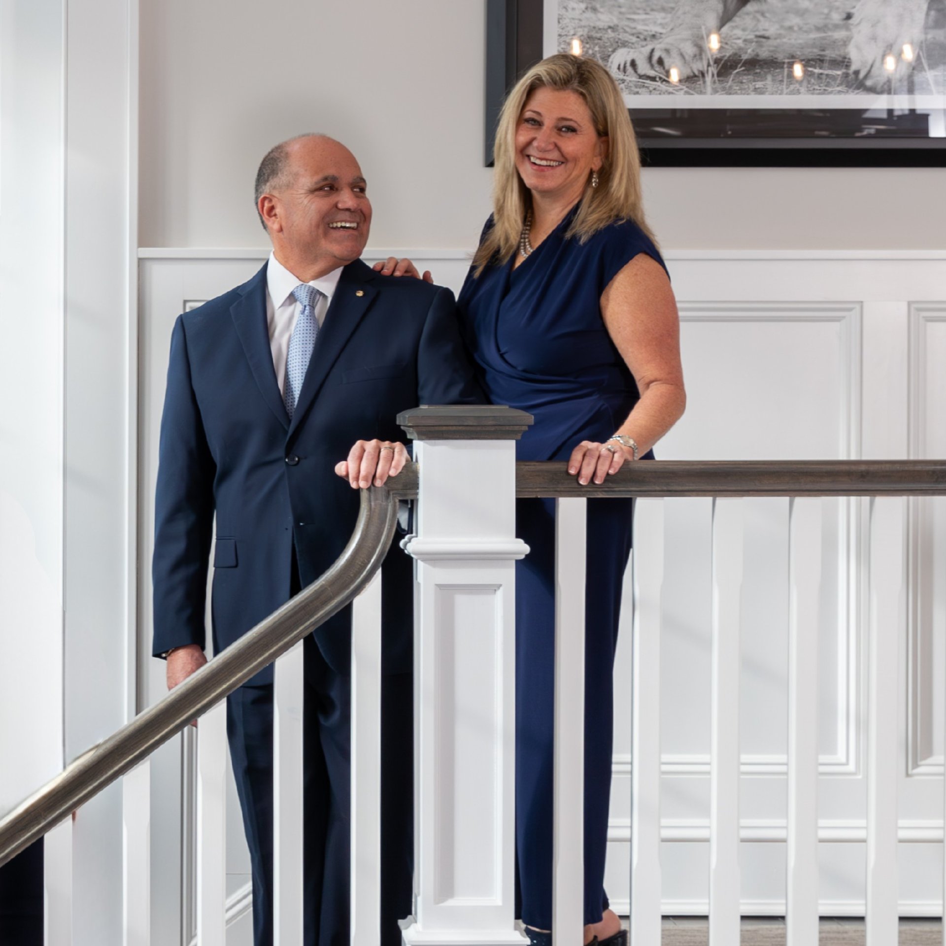 Two real estate professionals pose beside a staircase railing, the woman in a navy dress and man in a navy suit.