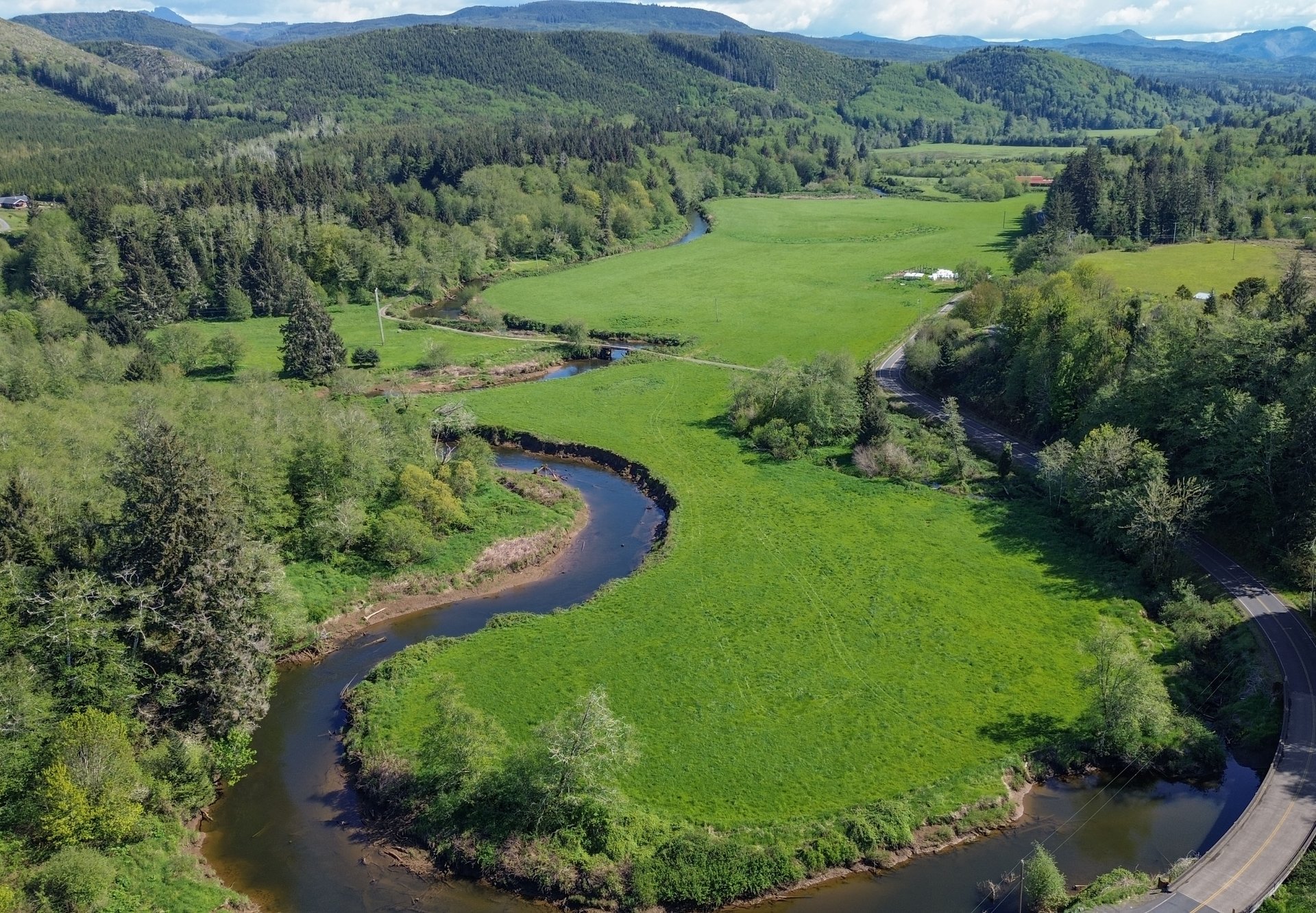 Aerial view of green pastureland with a winding creek, forested hills, and a road along the edge.
