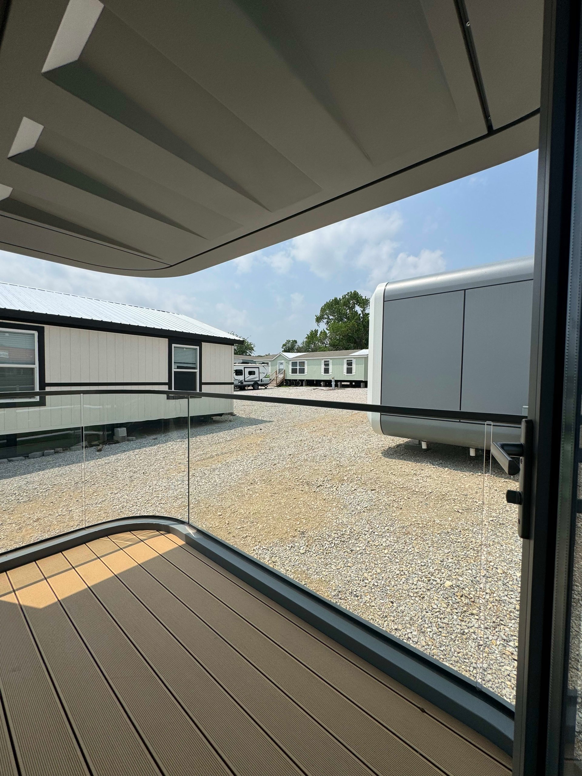 Covered tiny-home deck with brown plank flooring and glass railing overlooking a gravel lot and neighboring homes.