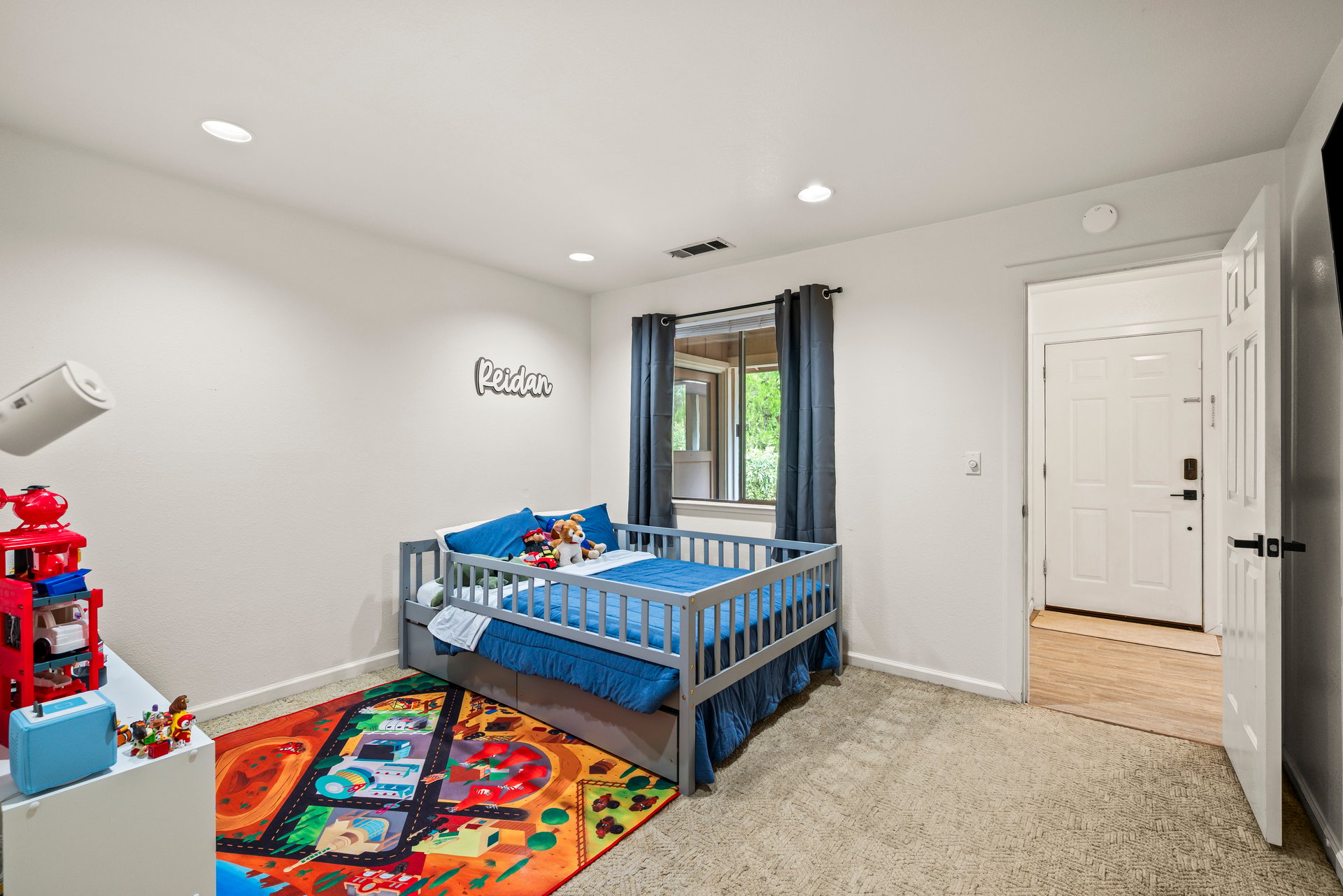Bedroom with a low gray bed, blue bedding, toys, and a colorful road-pattern rug.