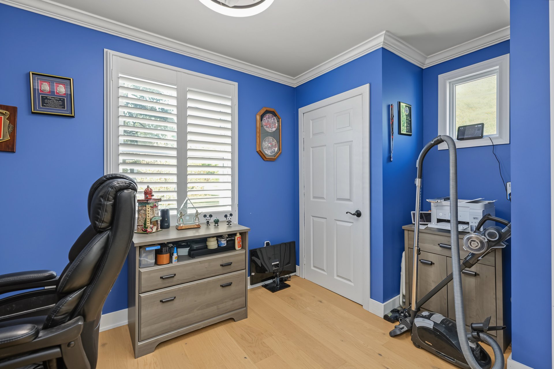 Home office with blue walls, white plantation shutters, and a leather chair beside exercise equipment.