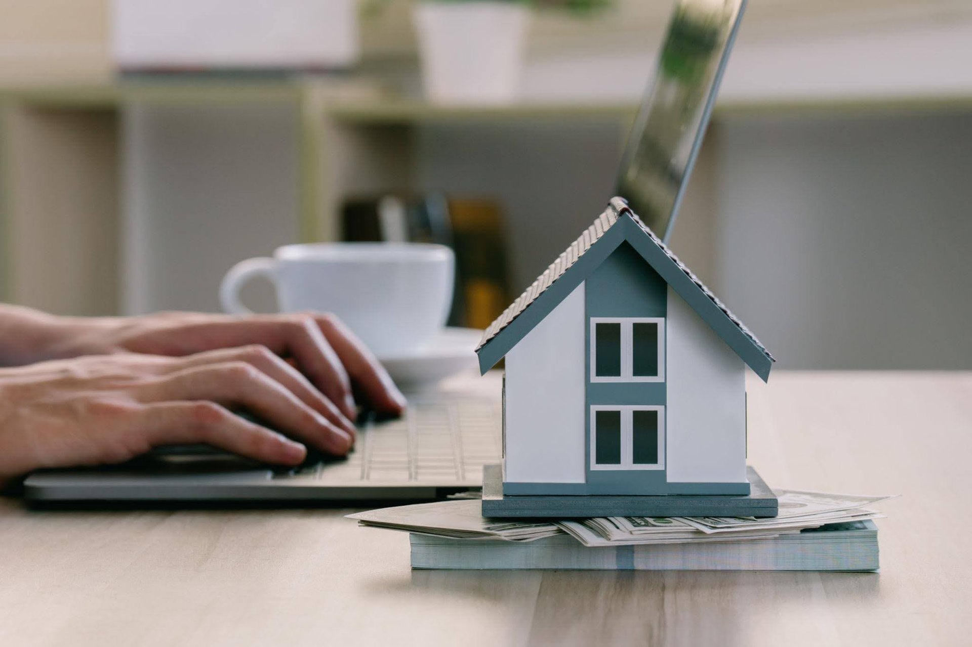 a-person-types-on-a-laptop-next-to-a-small-house-model-resting-on-dollar-bills-a-white-coffee-cup-is-blurred-in-the-background
