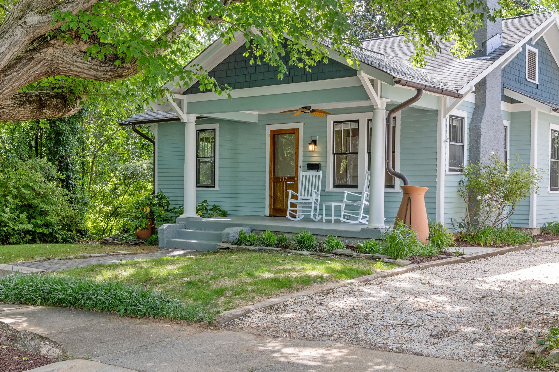 Teal house with a shaded front porch, white rocking chairs, and a large leafy tree.