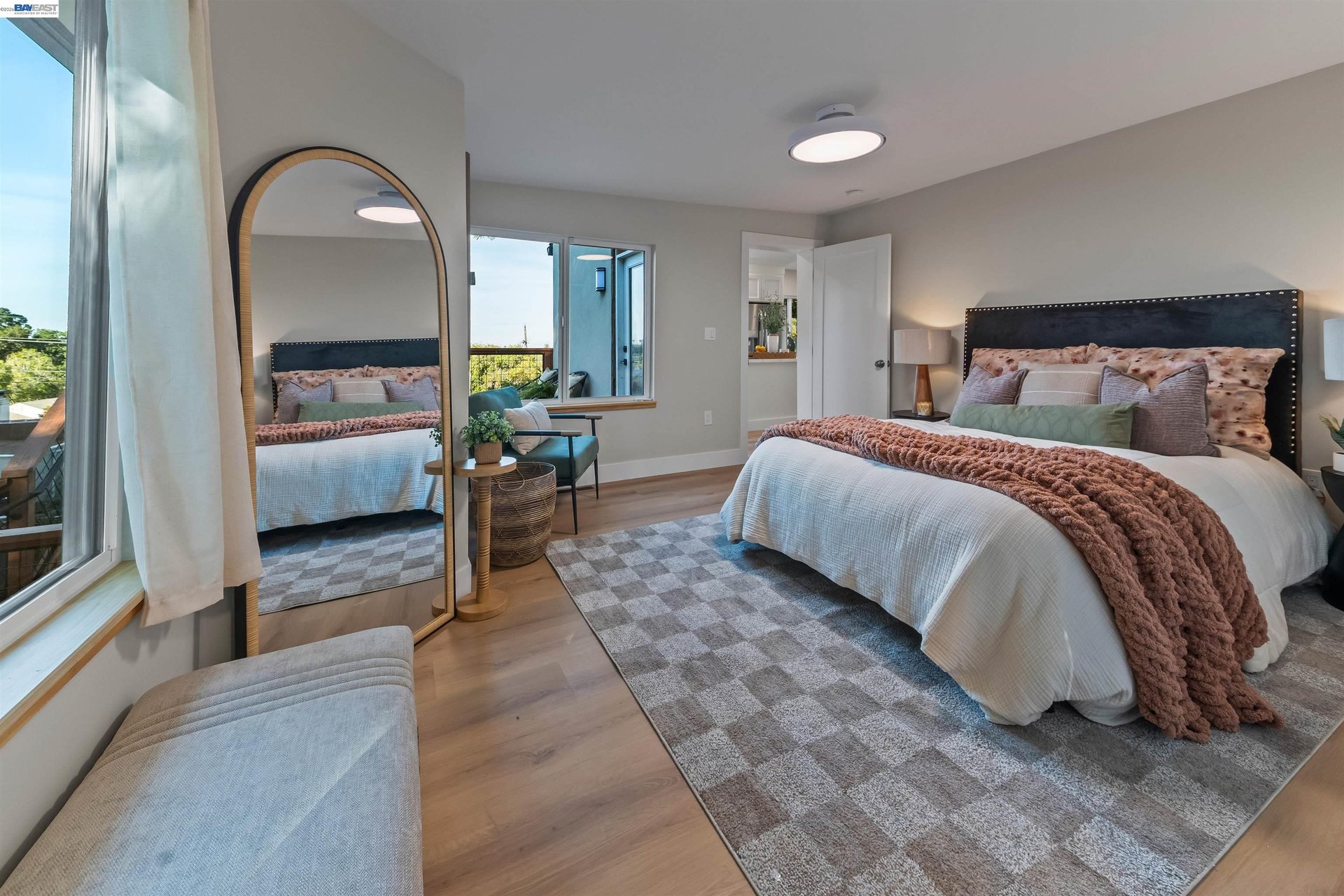 Staged bedroom with a large bed, arched mirror, patterned rug, and broad windows.