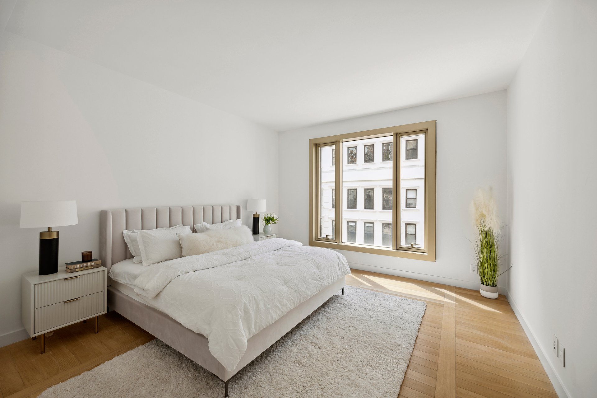 Staged bedroom with a large bed, hardwood floors, and a broad window overlooking a neighboring building.