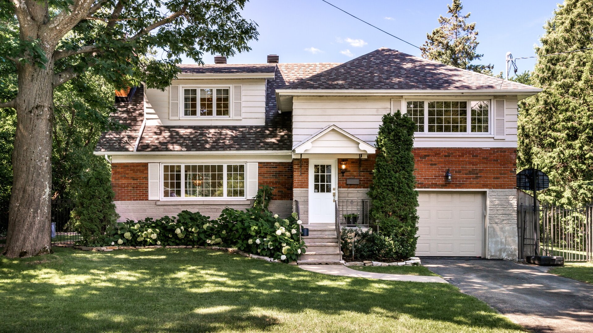 Two-story brick-and-siding house with an attached garage, landscaped shrubs, and mature trees.