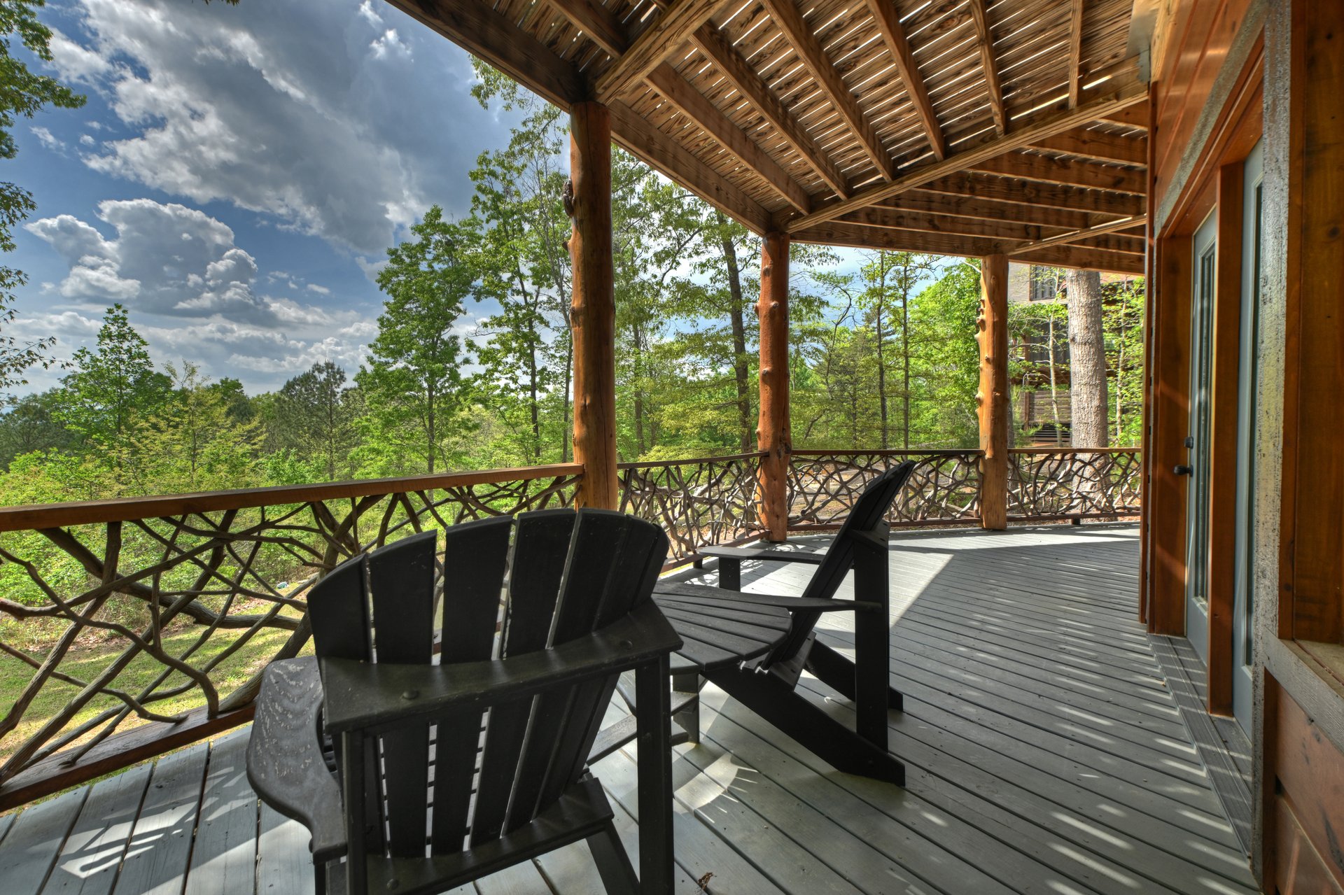Covered timber porch with Adirondack chairs and rustic branch railings overlooking trees.