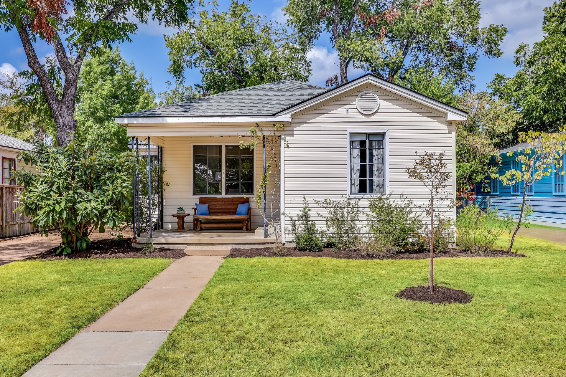 Cream-sided single-story house with a covered porch, wooden bench, and walkway through a green lawn.