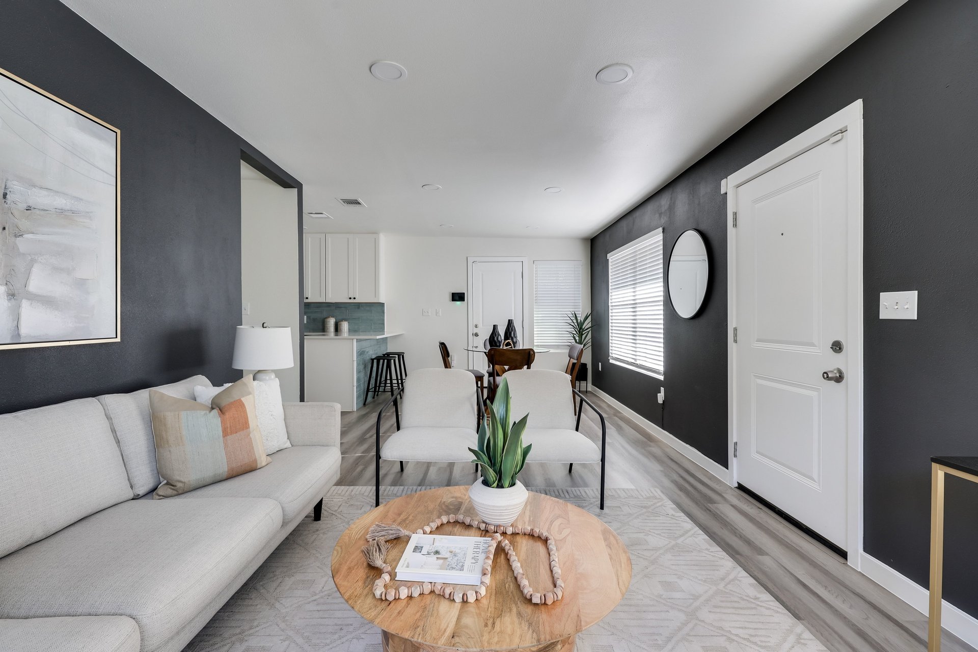 Staged living room with a light sectional, round wood coffee table, charcoal walls, and an open kitchen.