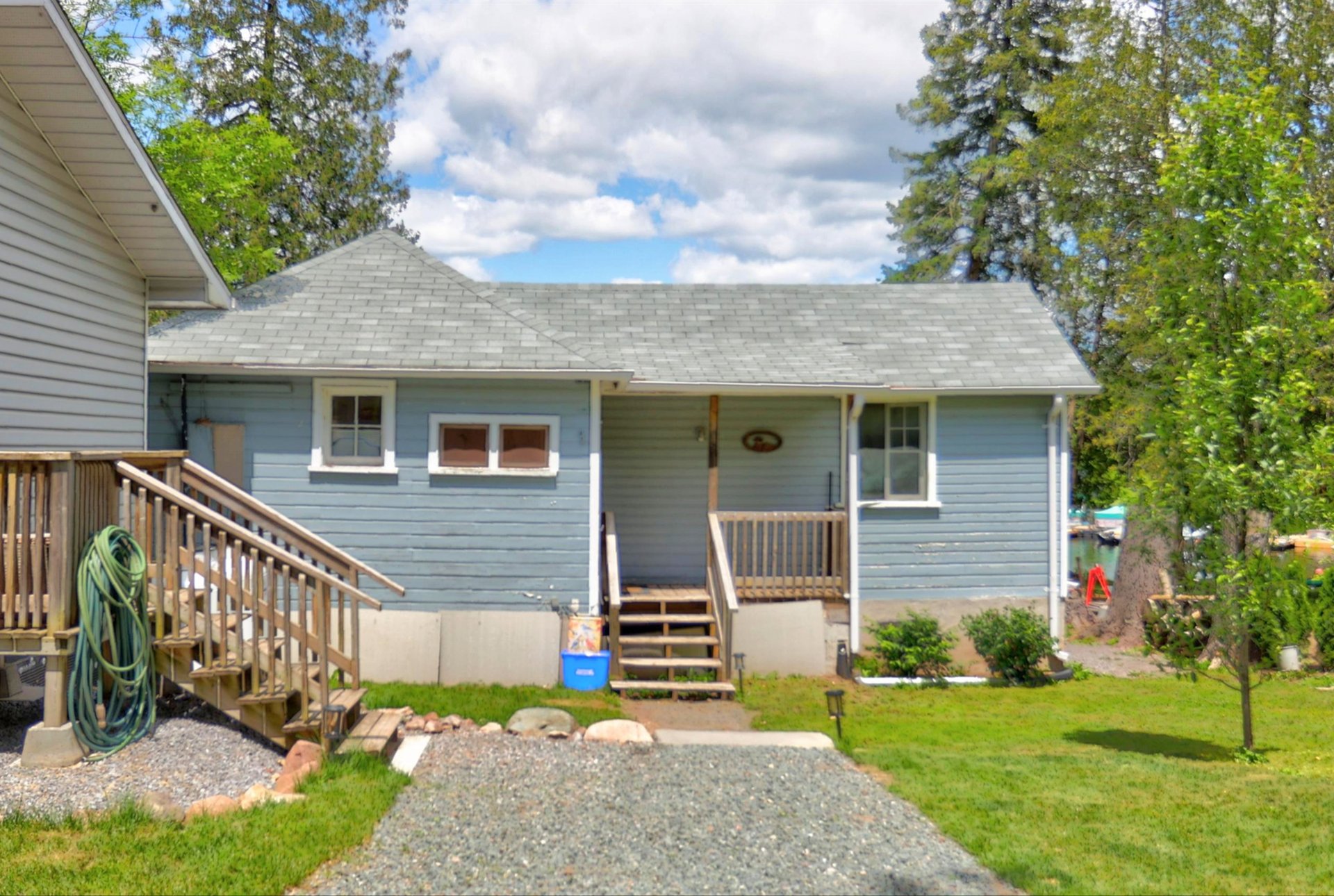 Blue cottage with a wooden front porch, gravel driveway, and a glimpse of water nearby.