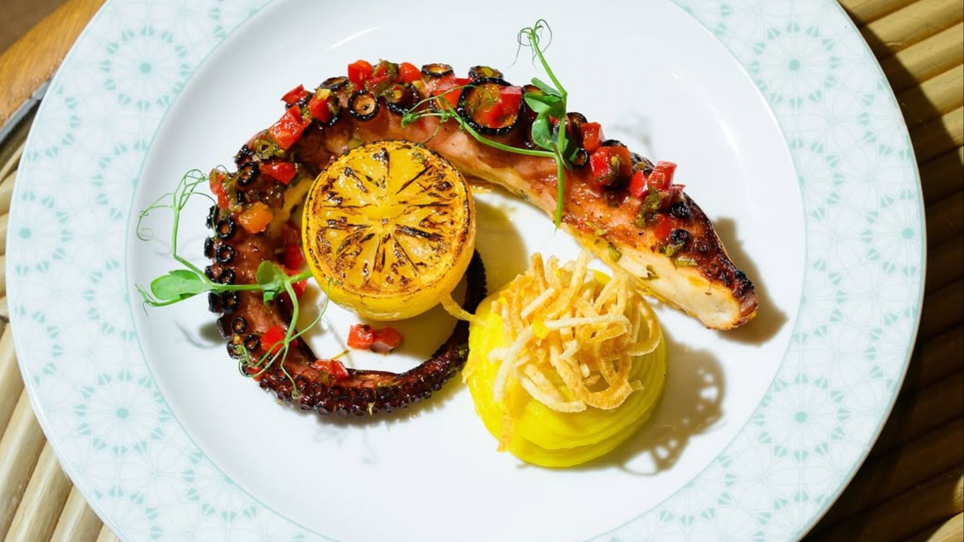 Charred octopus with grilled lemon half and pasta nest garnish on white plate with decorative rim
