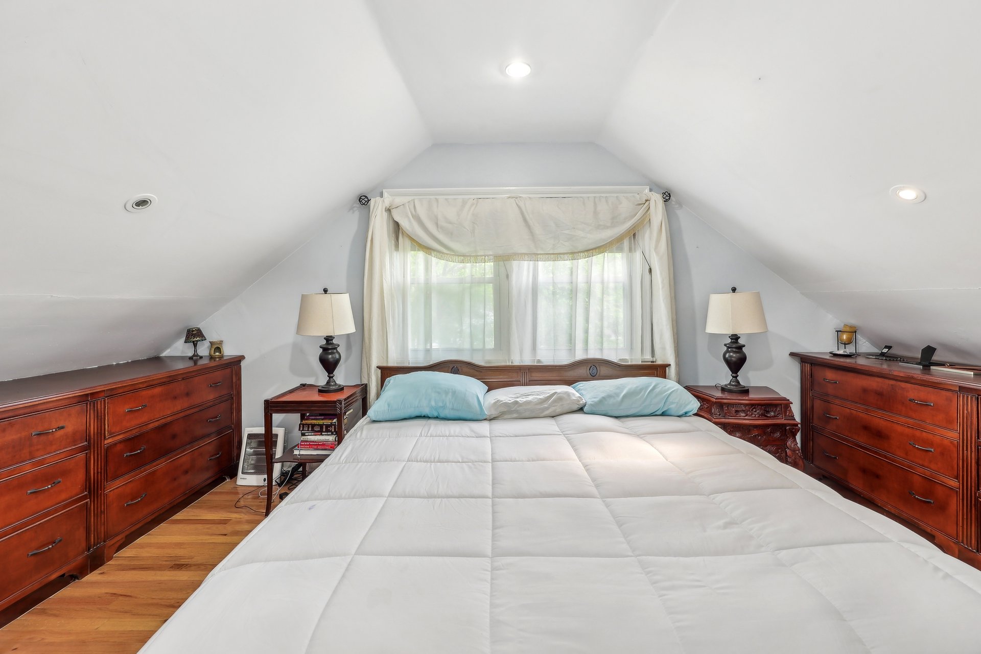 Attic bedroom with a large bed, wooden dressers, and a curtained window beneath a sloped ceiling.