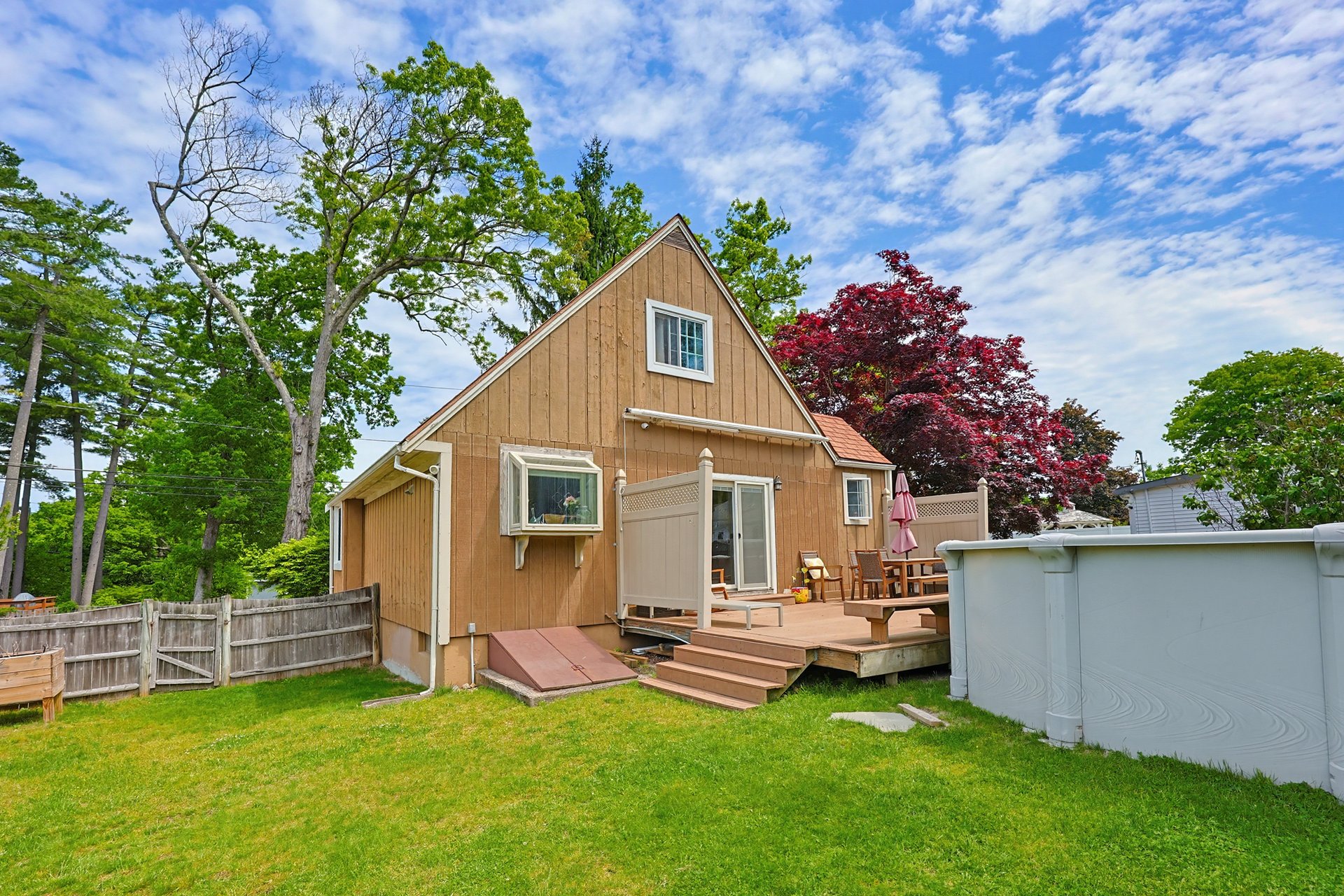 Tan house with a raised deck and above-ground pool in a fenced, tree-lined backyard.