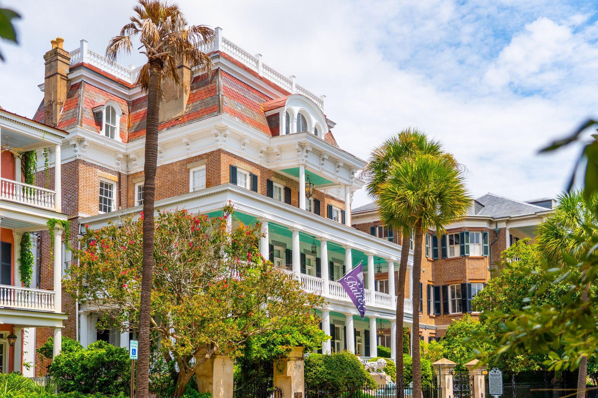 Historic brick Charleston house with a red mansard roof, columned porches, and palm trees.