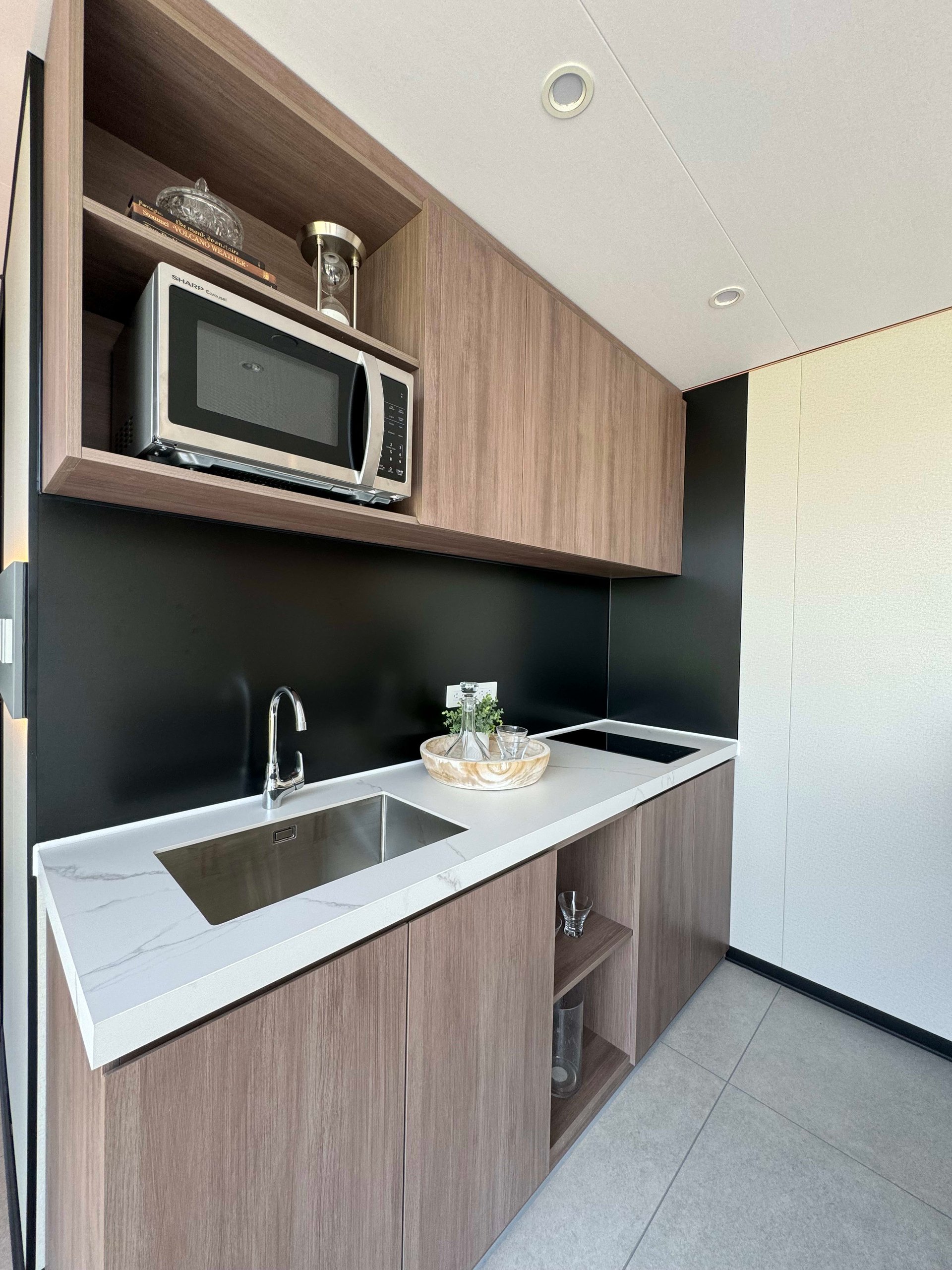 Compact tiny-home kitchen with wood cabinetry, a white veined counter, stainless sink, and built-in microwave.