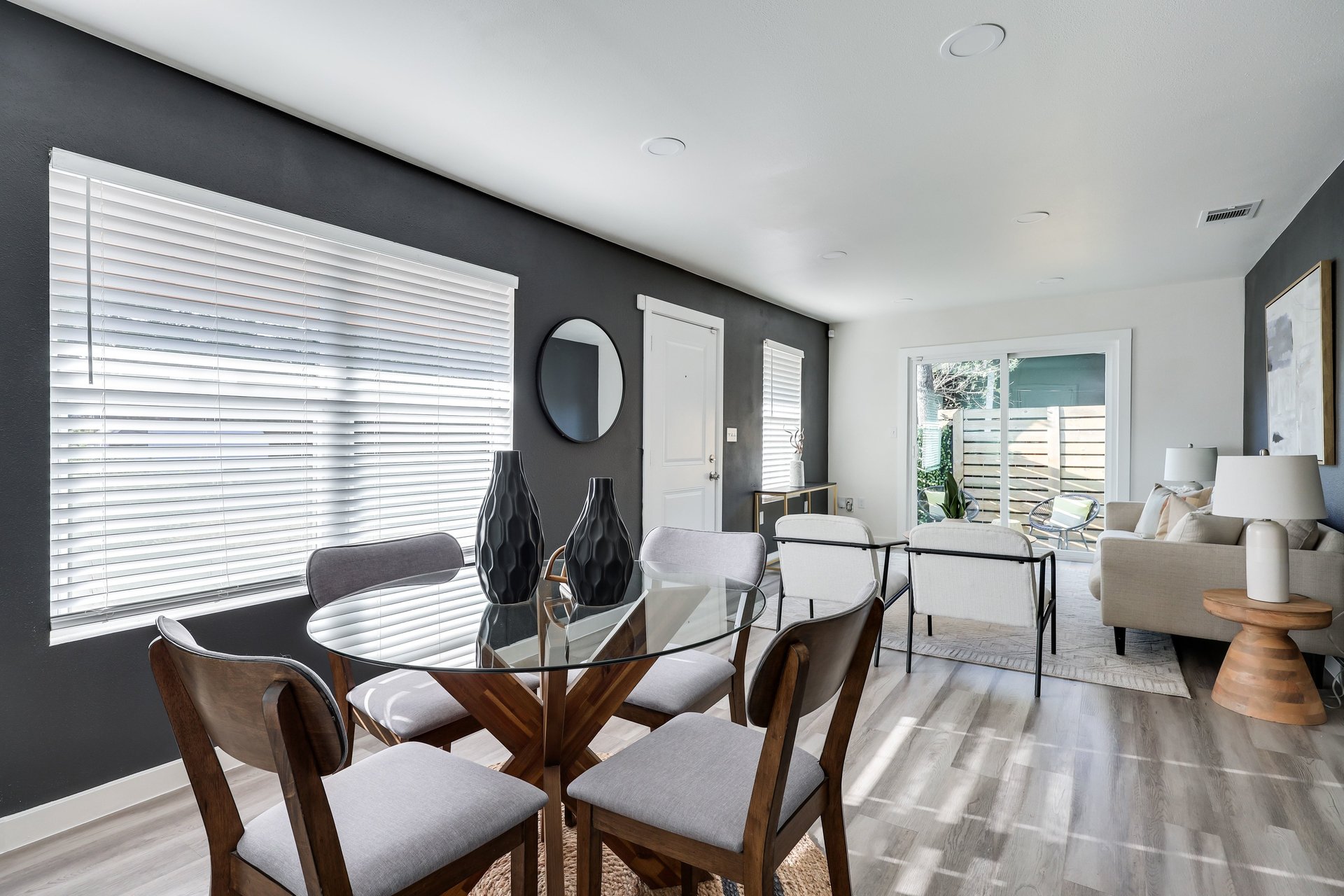 Dining and living area with a glass-top table, charcoal accent wall, and sliding doors to a fenced patio.
